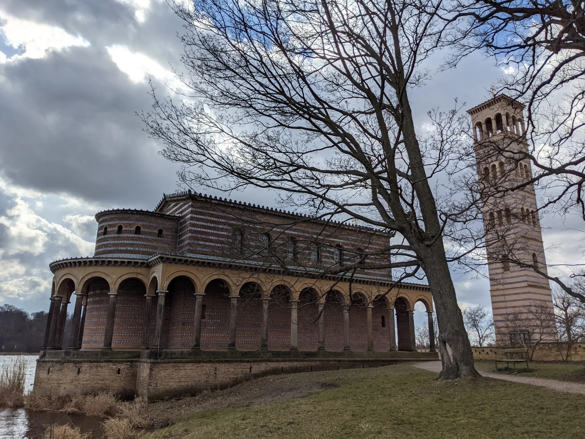 Kirche aus gestreiftem Backstein mit umlaufendem Arkadengang direkt am Wasser, rechts der freistehende Glockenturm, davor ein kahler Baum