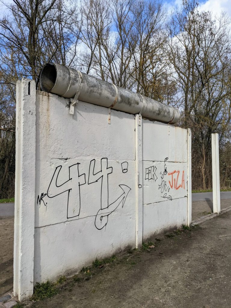 Weiß gestrichenes Stück Grenzmauer aus Betonplatten mit einem Rohr als Mauerkrone, darauf Graffiti