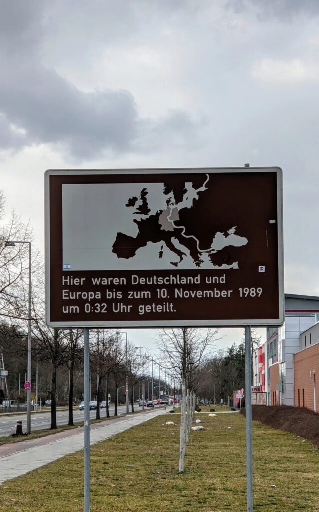 Braunes Hinweisschild mit Europakarte und dem Text „Hier waren Deutschland und Europa bis zum 10. November 1989 um 0:32 Uhr geteilt“ an einer breiten Straße