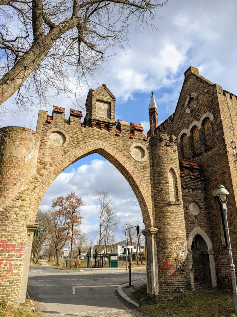 Neogotisches Tor aus Backstein mit Spitzbogen, Zinnen und kleinen Türmen, dahinter eine Straßenkreuzung