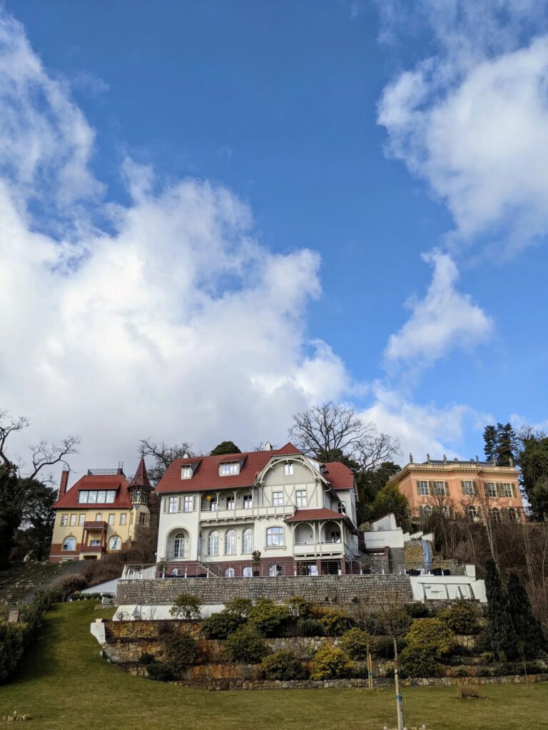 Drei Villen mit roten Dächern und Terrassen an einem grünen Hang unter blauem Himmel mit Wolken