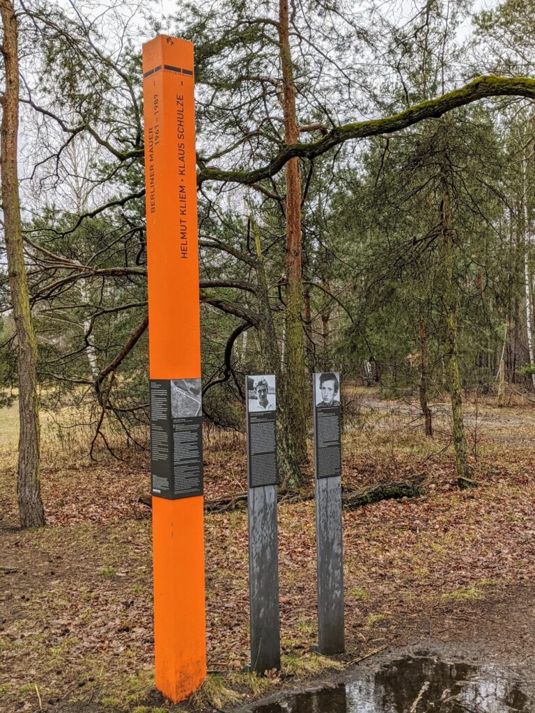 Orangefarbene Stele mit den Namen Helmut Kliem und Klaus Schulze, daneben zwei schmale Tafeln mit Porträtfotos im Kiefernwald