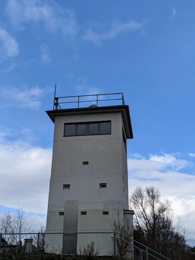 Grauer, viereckiger Wachturm mit Fensterband und Geländer auf dem Dach vor blauem Himmel