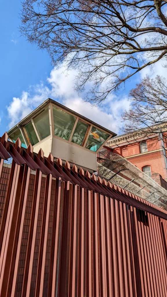 Wachturm hinter rotem Zaun mit Stacheldraht an der Justizvollzugsanstalt Moabit, dahinter Backsteinbau