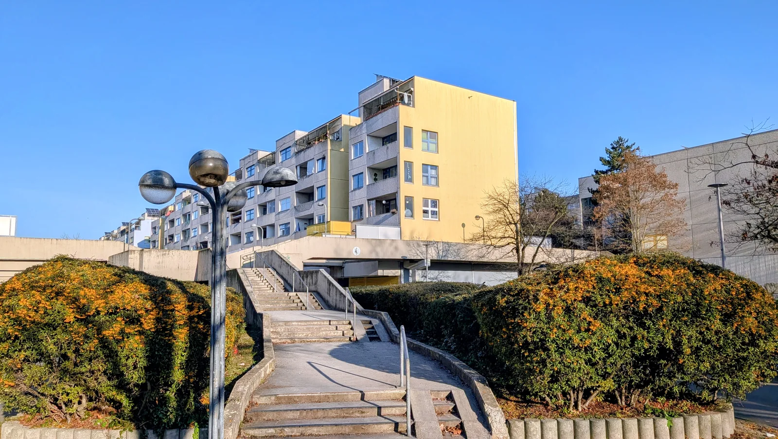 Gelb verputzter Wohnblock der High-Deck-Siedlung mit Loggien über einer Betonzeile, davor Treppen, Kugelleuchte und Büsche mit orangen Beeren
