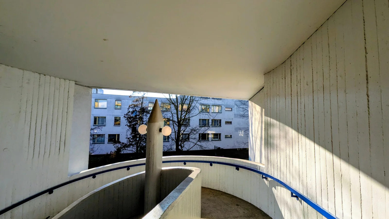 Rampe mit blauem Geländer und Betonpfeiler zwischen Sichtbetonwänden in der High-Deck-Siedlung, im Durchblick ein hellblau gestrichener Wohnblock
