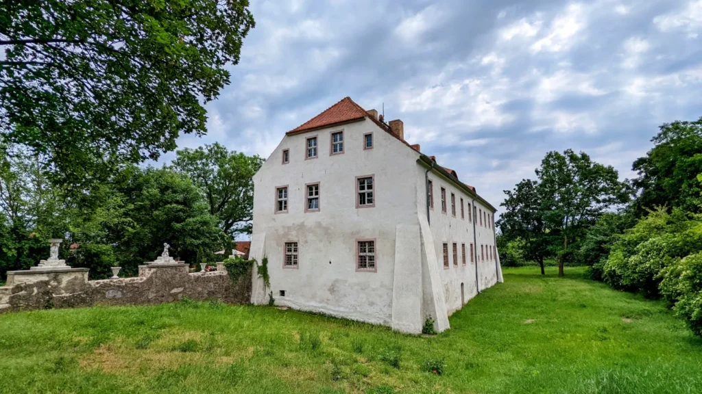 Schloss Neuenhagen, ein weiß verputztes Festes Haus mit rotem Dach und Strebepfeilern an der Ecke, davor eine Wiese
