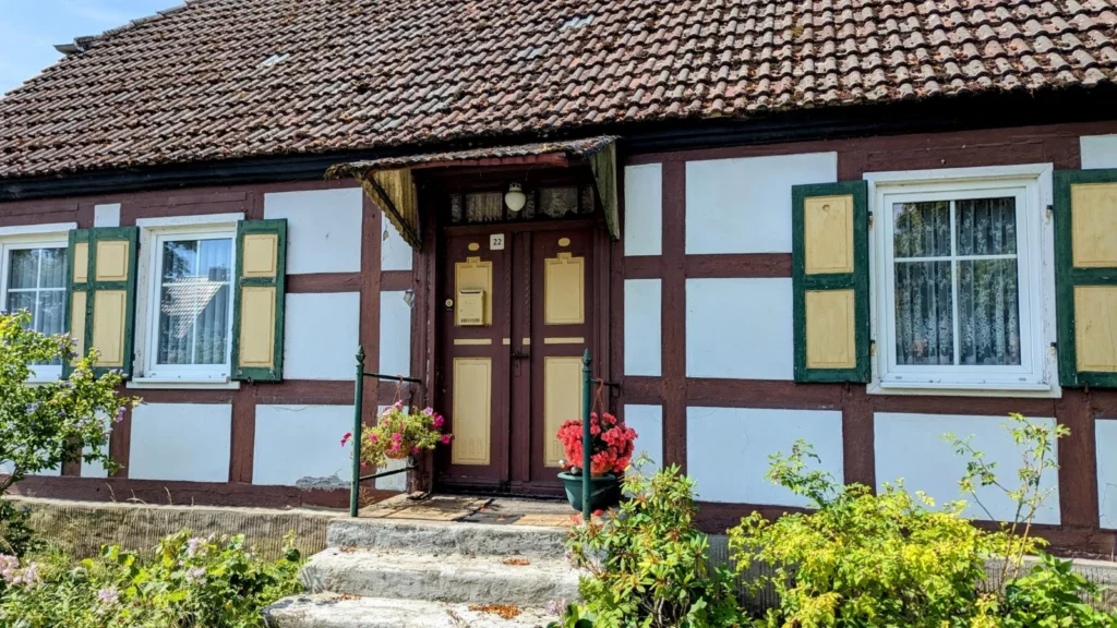 Eingeschossiges Fachwerkhaus mit weißen Gefachen, grün-gelben Fensterläden und Blumen an der Eingangstreppe in Rathsdorf