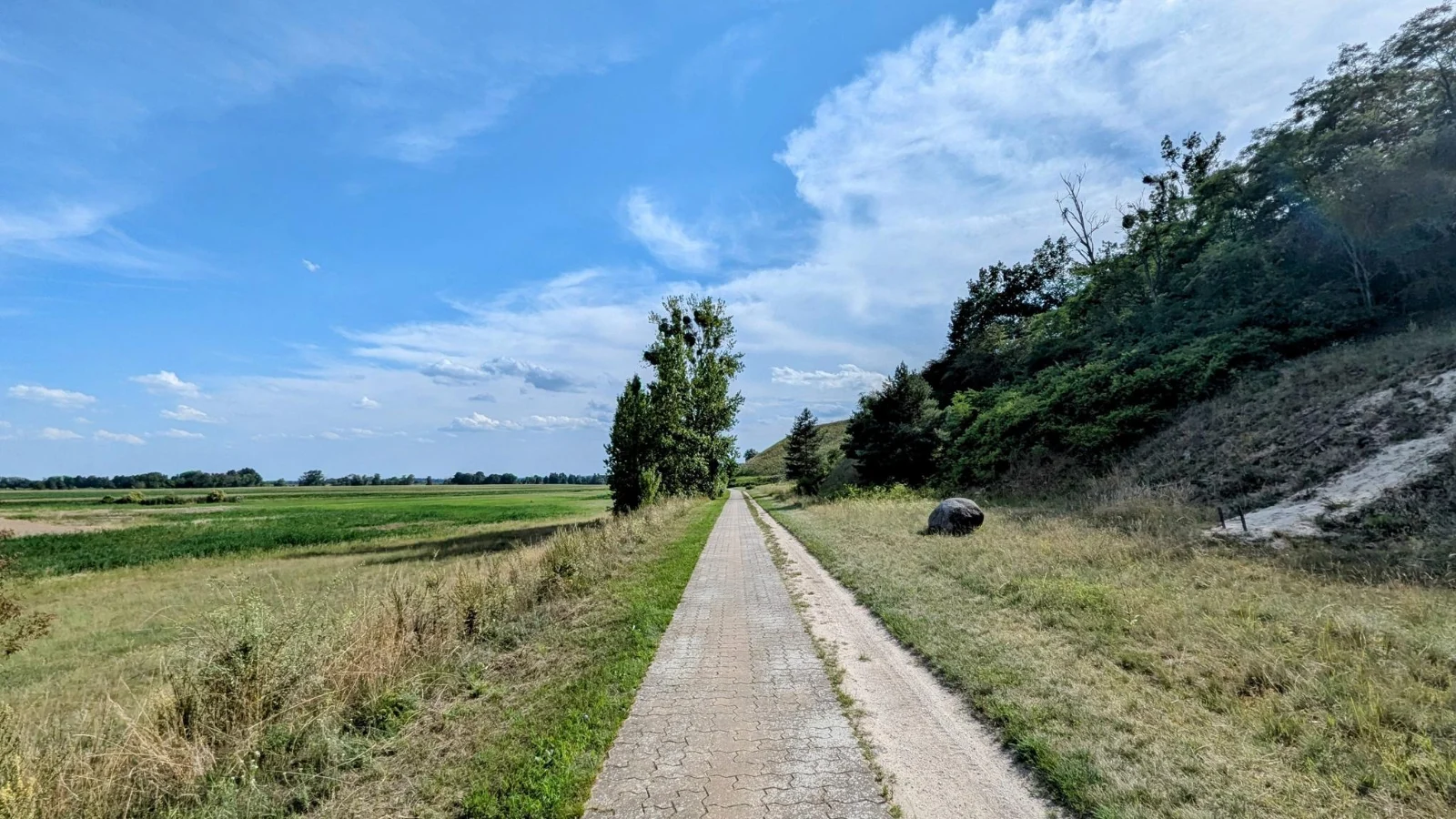 Gepflasterter Weg am Fuß eines bewachsenen Hangs mit offener Sandstelle, links die weite Ebene des Oderbruchs