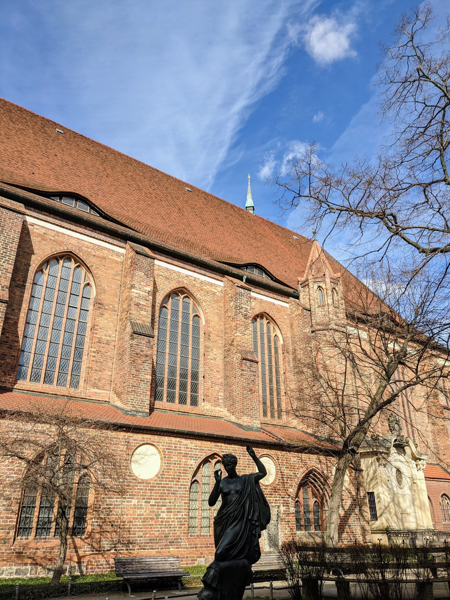 Seitenansicht einer Backsteinkirche mit hohen Spitzbogenfenstern und steilem Ziegeldach, davor die dunkle Figur einer Frau in wallendem Gewand, kahle Bäume, blauer Himmel