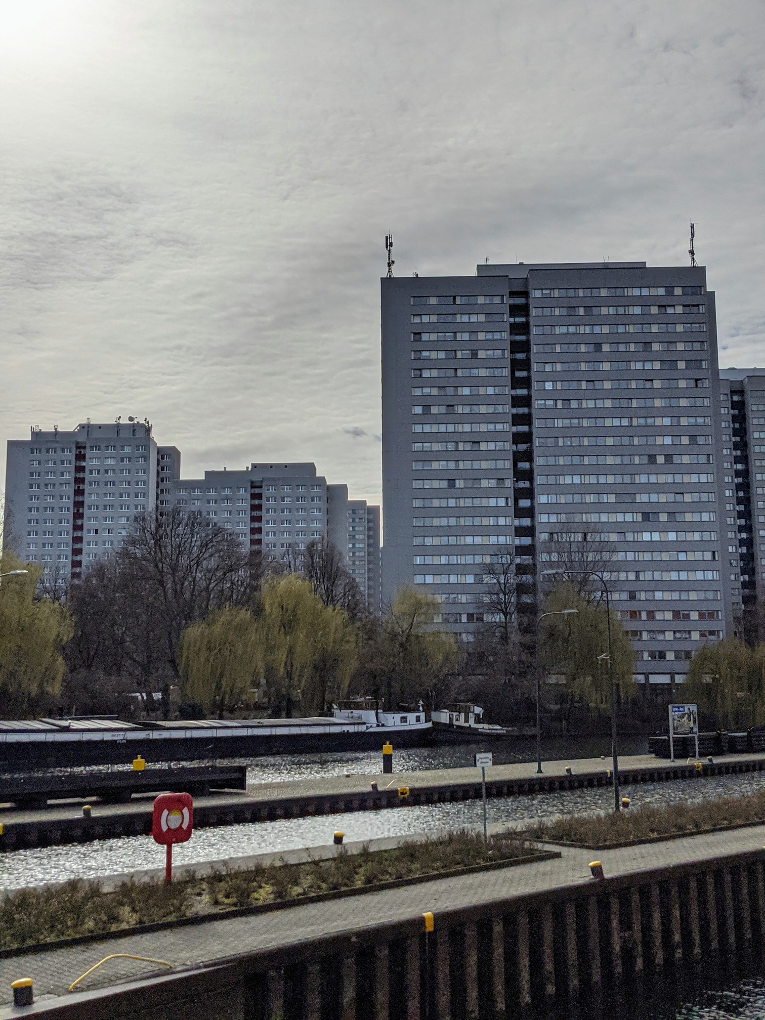 Graue Plattenbau-Hochhäuser hinter kahlen Bäumen und Trauerweiden, davor ein Gewässer mit Kaimauern und Lastkähnen, wolkiger Himmel