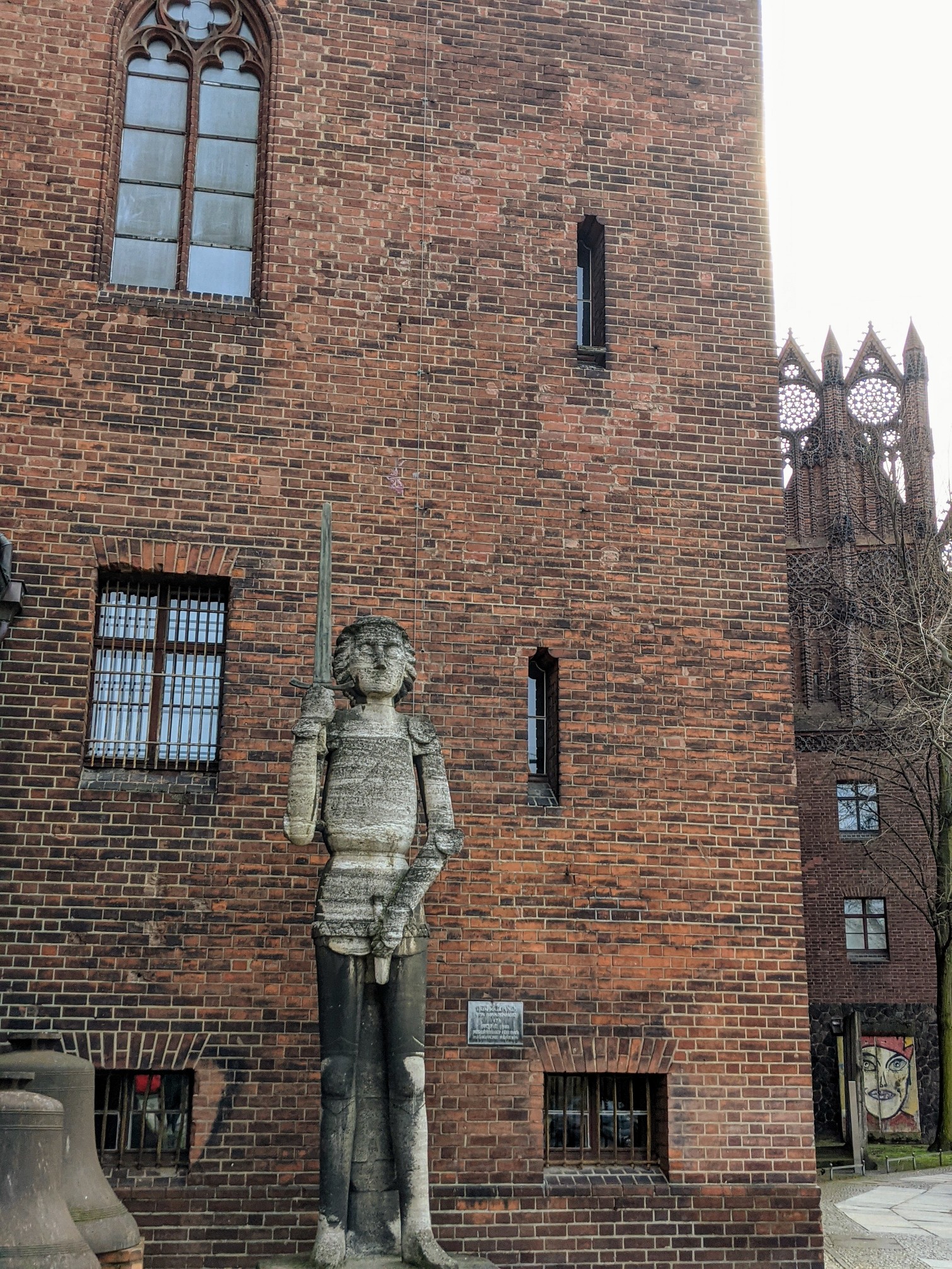 Steinerne Ritterfigur mit erhobenem Schwert vor einer hohen Backsteinwand, links unten zwei Glocken, rechts dahinter ein Backsteingiebel mit Rosetten und ein bemaltes Mauersegment