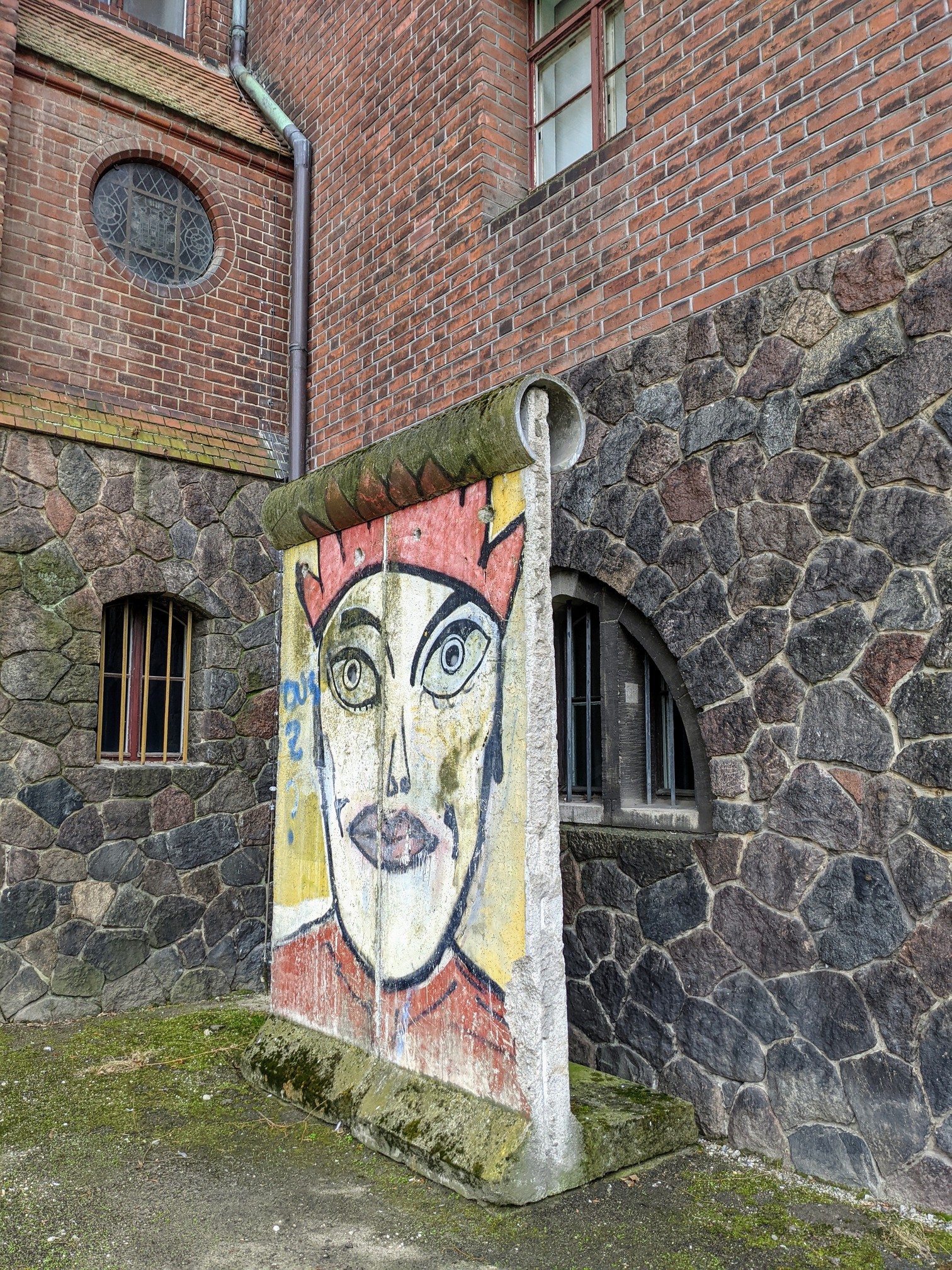 Bemaltes Betonsegment mit Rohraufsatz, darauf ein Gesicht mit roter Zackenkrone, vor einer Wand aus Backstein und Feldsteinen