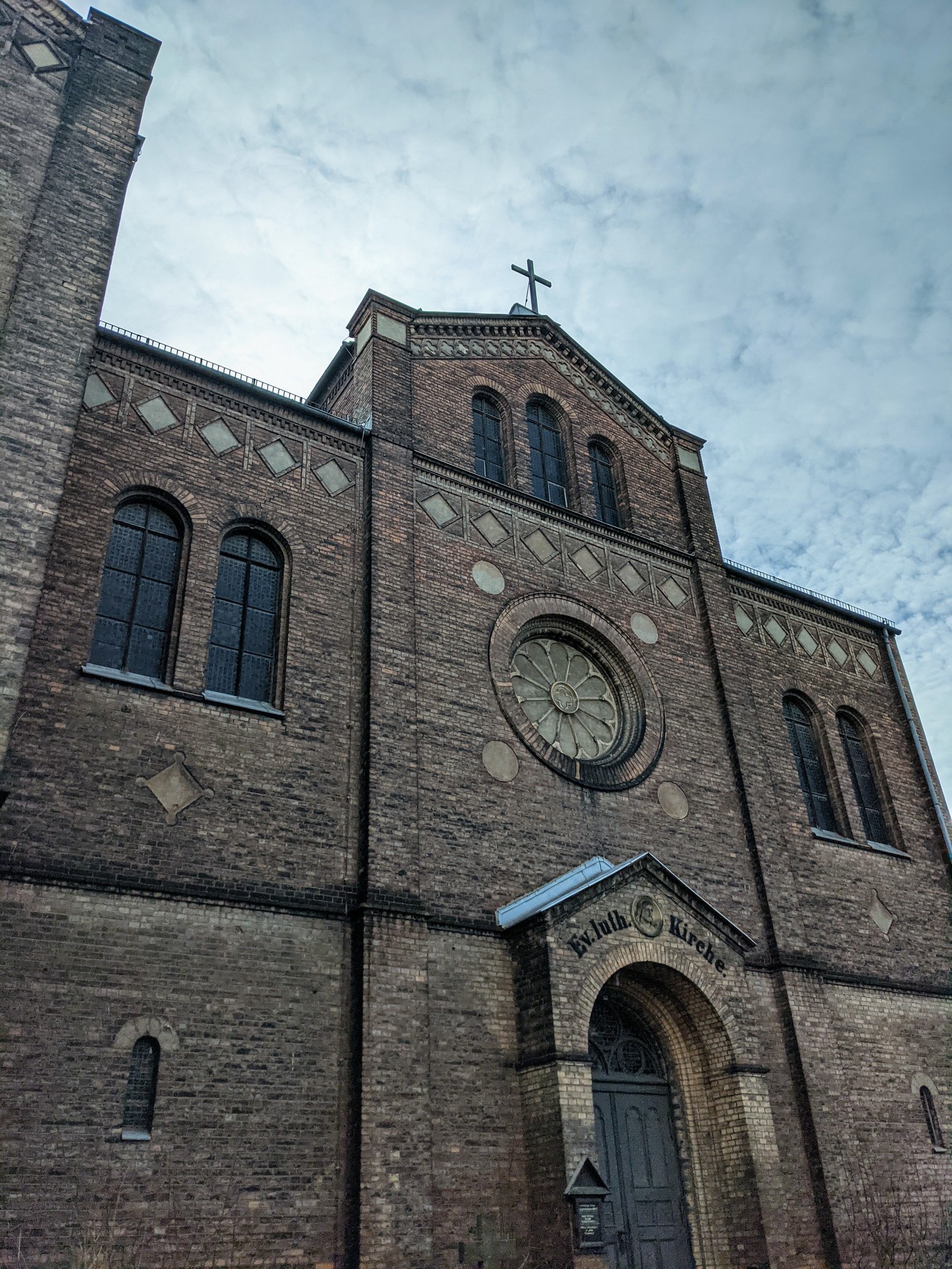 Dunkle Backsteinfassade mit Rosettenfenster, Rundbogenportal und Kreuz auf dem Giebel, über dem Portal die Inschrift „Ev. luth. Kirche.“