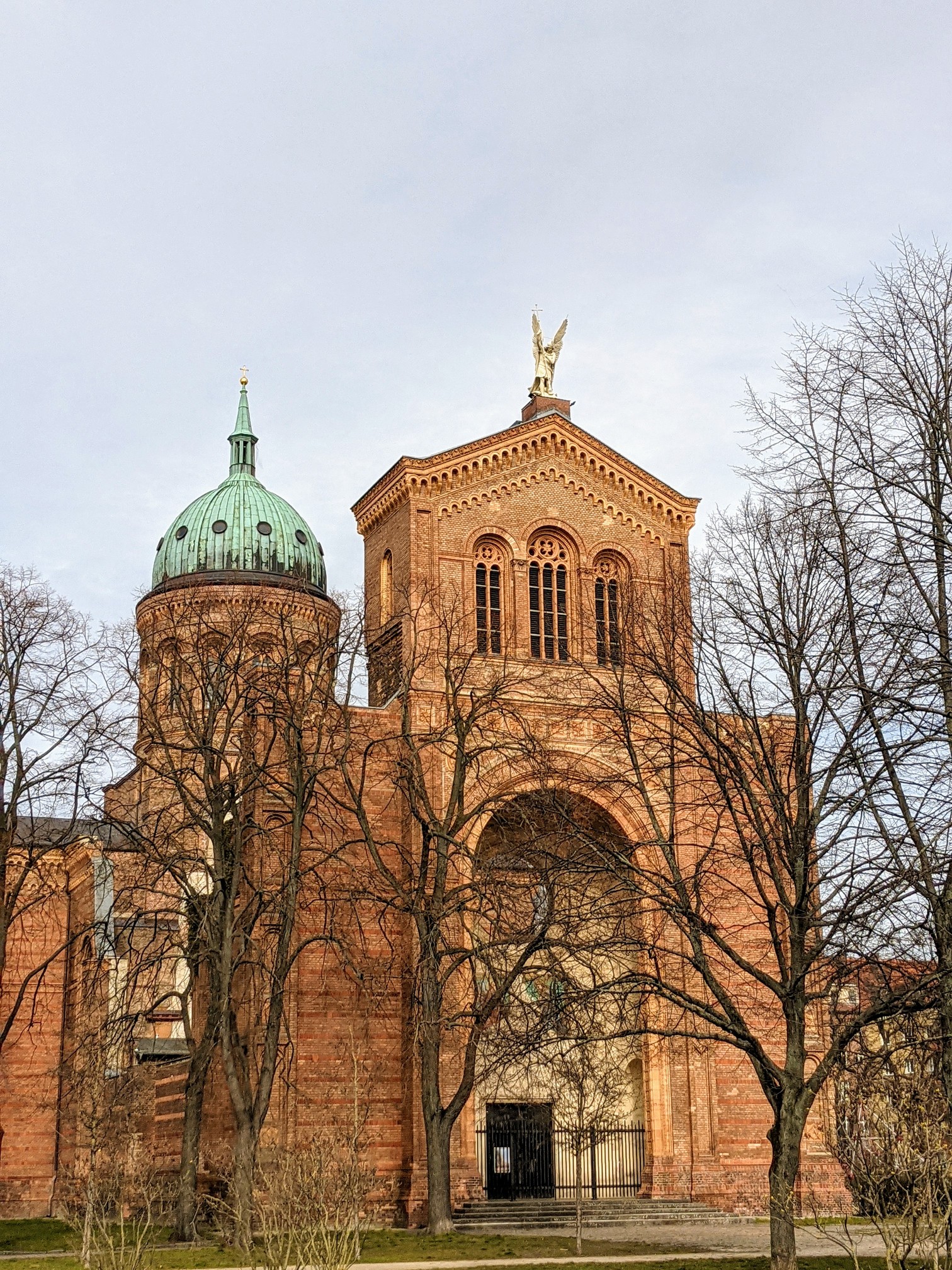 Rote Backsteinkirche mit grüner Kupferkuppel links und einem Turm mit goldener Engelsfigur auf dem Giebel, davor kahle Bäume