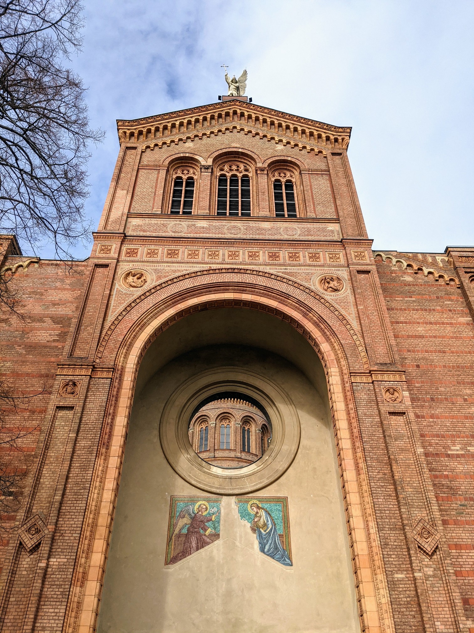 Hohe Backsteinfassade mit großem Rundbogen, darin ein Rundfenster und darunter ein Mosaik mit zwei Figuren, oben auf dem Giebel eine goldene Engelsfigur