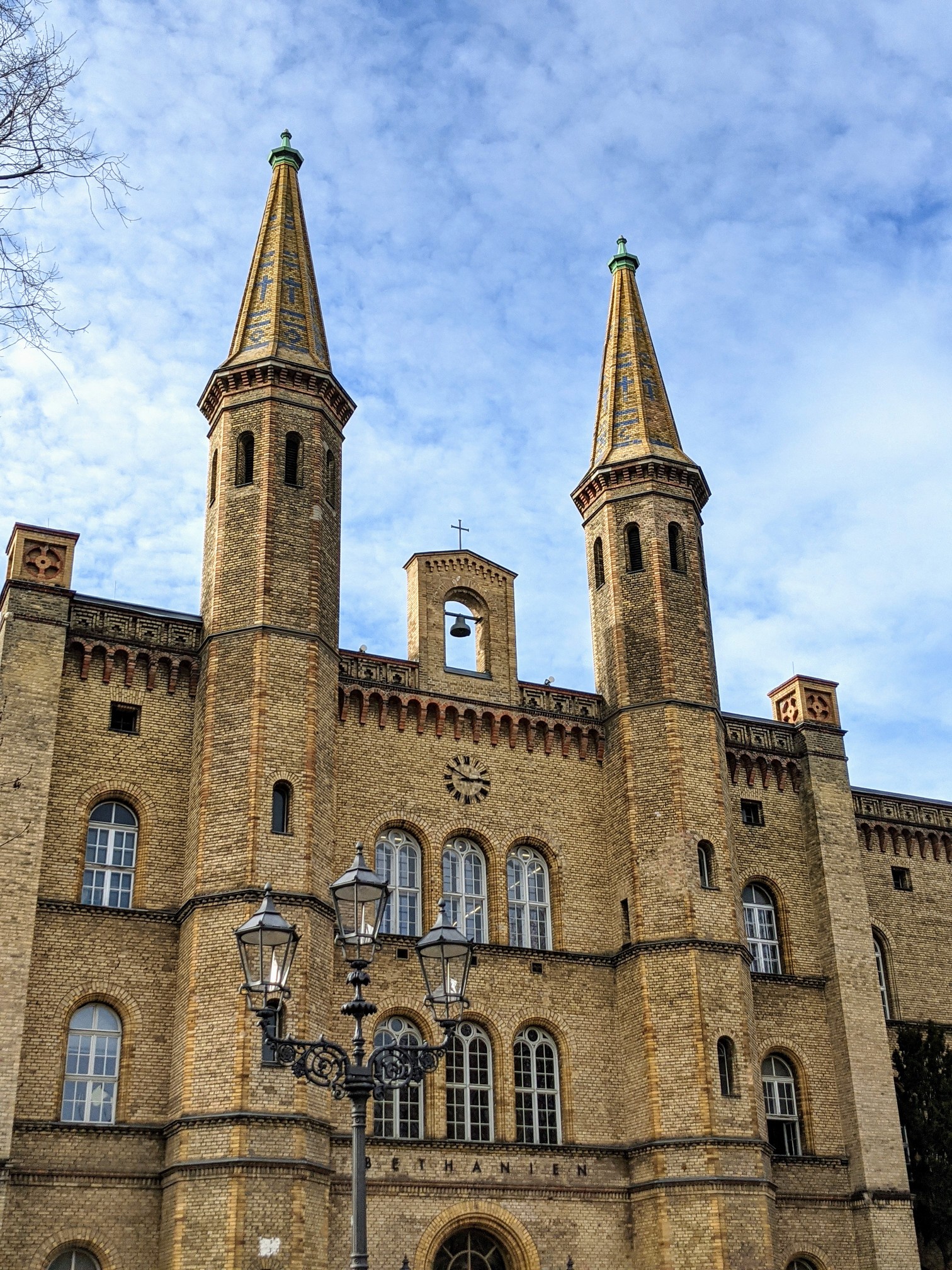 Gelbe Backsteinfassade mit zwei schlanken Türmen mit gemusterten Spitzen, dazwischen ein Dachreiter mit Glocke, davor eine Laterne, am Gebäude der Schriftzug BETHANIEN