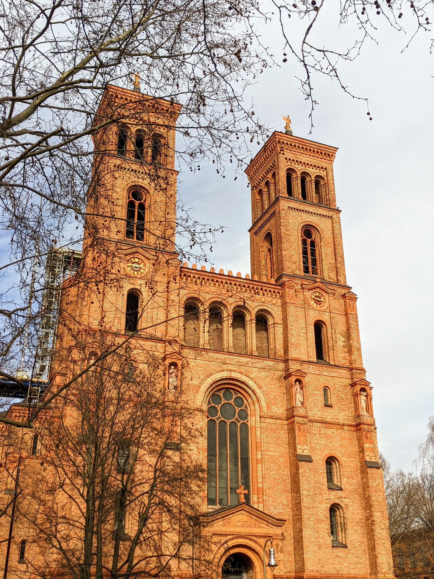Orangerote Backsteinkirche mit zwei Türmen und Uhren, teils hinter kahlen Ästen, links ein Gerüst