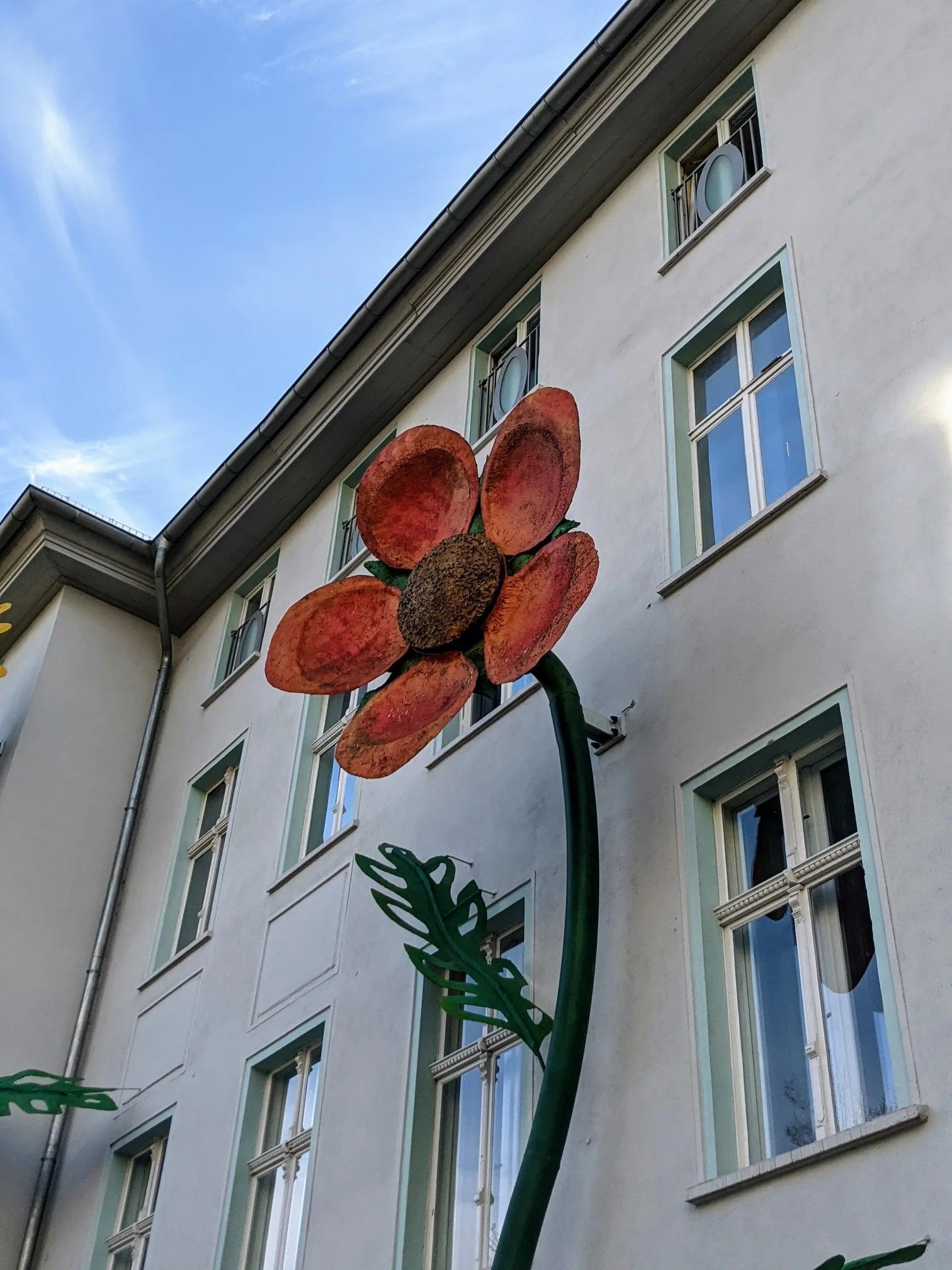 Große orangerote Blumenskulptur mit braunem Zentrum, grünem Stiel und Blatt an einer weißen Hauswand mit mintgrünen Fensterrahmen
