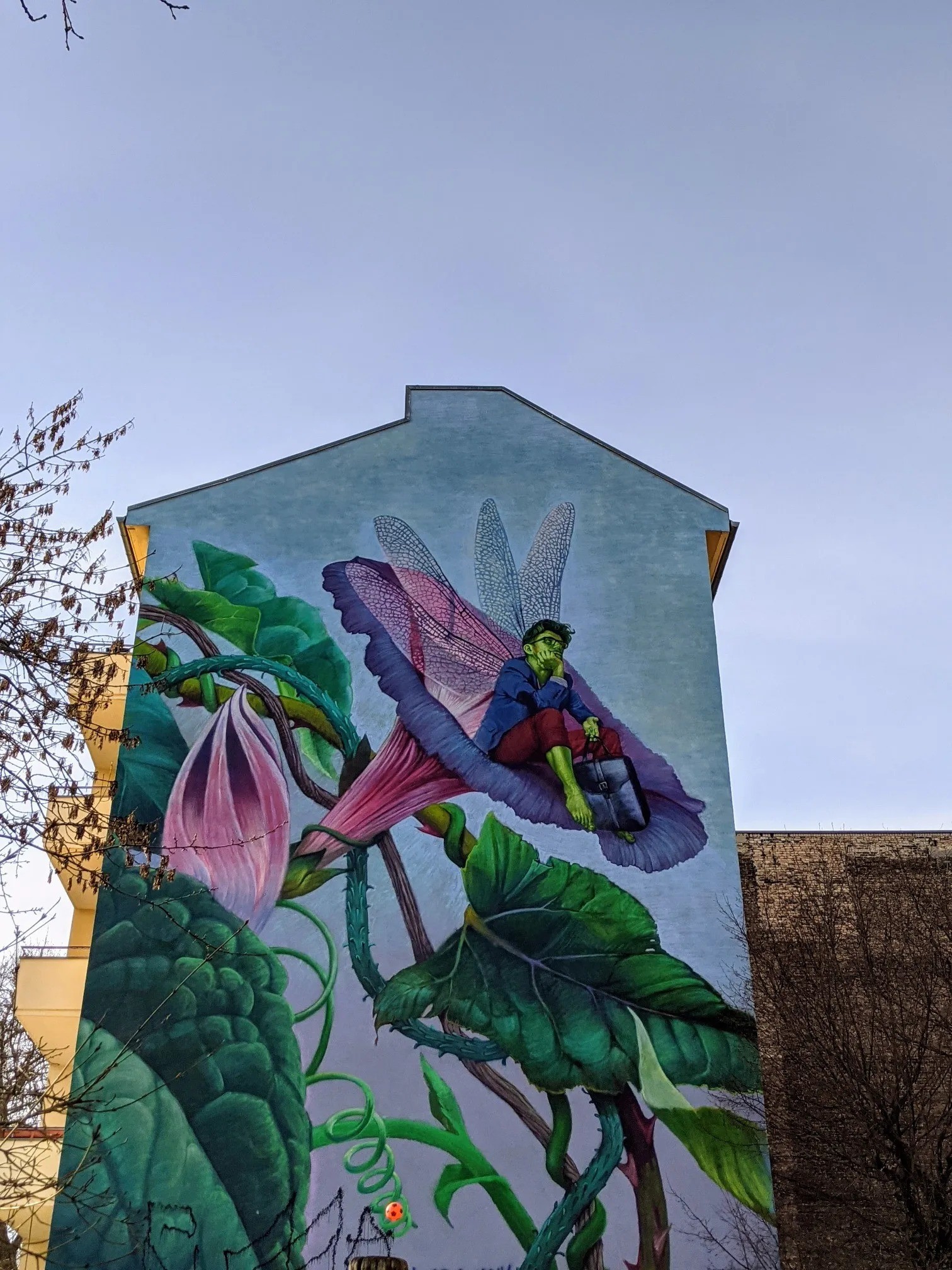Wandbild an einer Giebelwand: Ein Wesen mit Libellenflügeln, blauem Sakko und roter Hose sitzt mit Aktentasche in einer riesigen Blüte, daneben Knospe und große Blätter