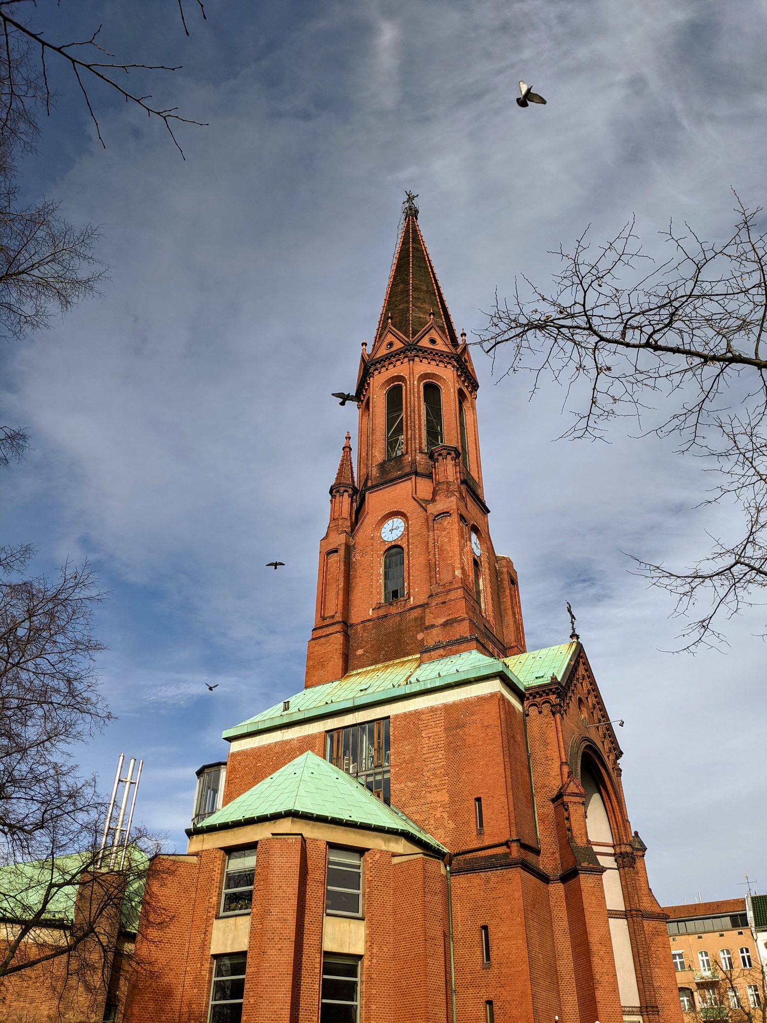 Rote Backsteinkirche mit spitzem Turm mit Uhr, grünen Kupferdächern und modernem Anbau, Tauben im Flug