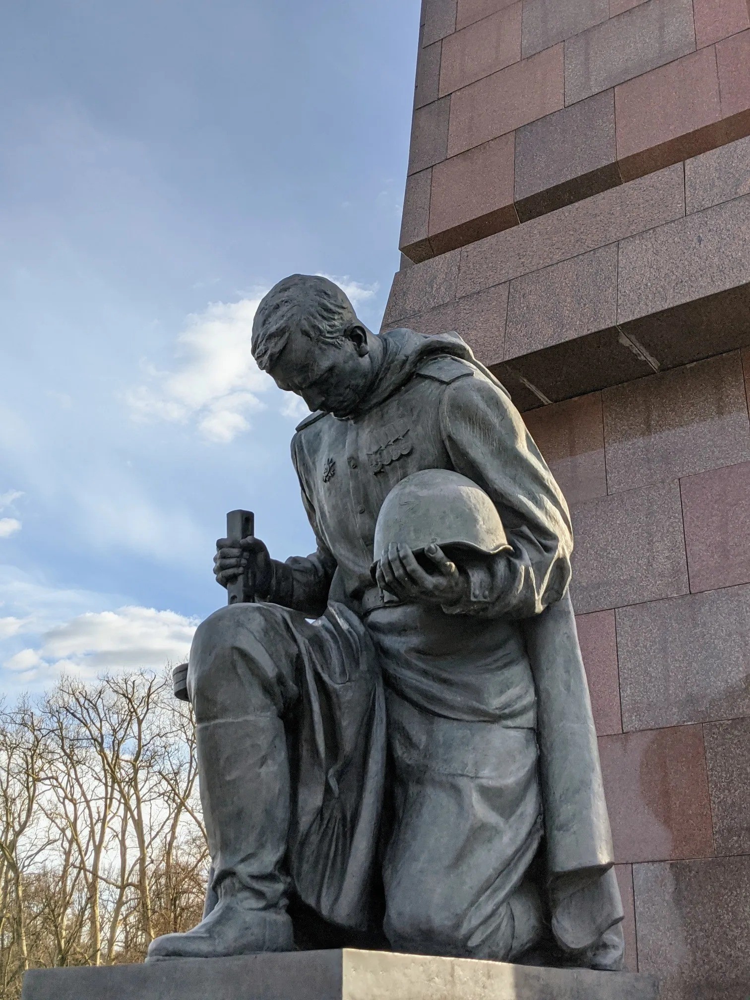 Dunkle Figur eines knienden Soldaten mit gesenktem Kopf, der einen Helm im Arm hält, vor einem Obelisken aus rötlichem Granit