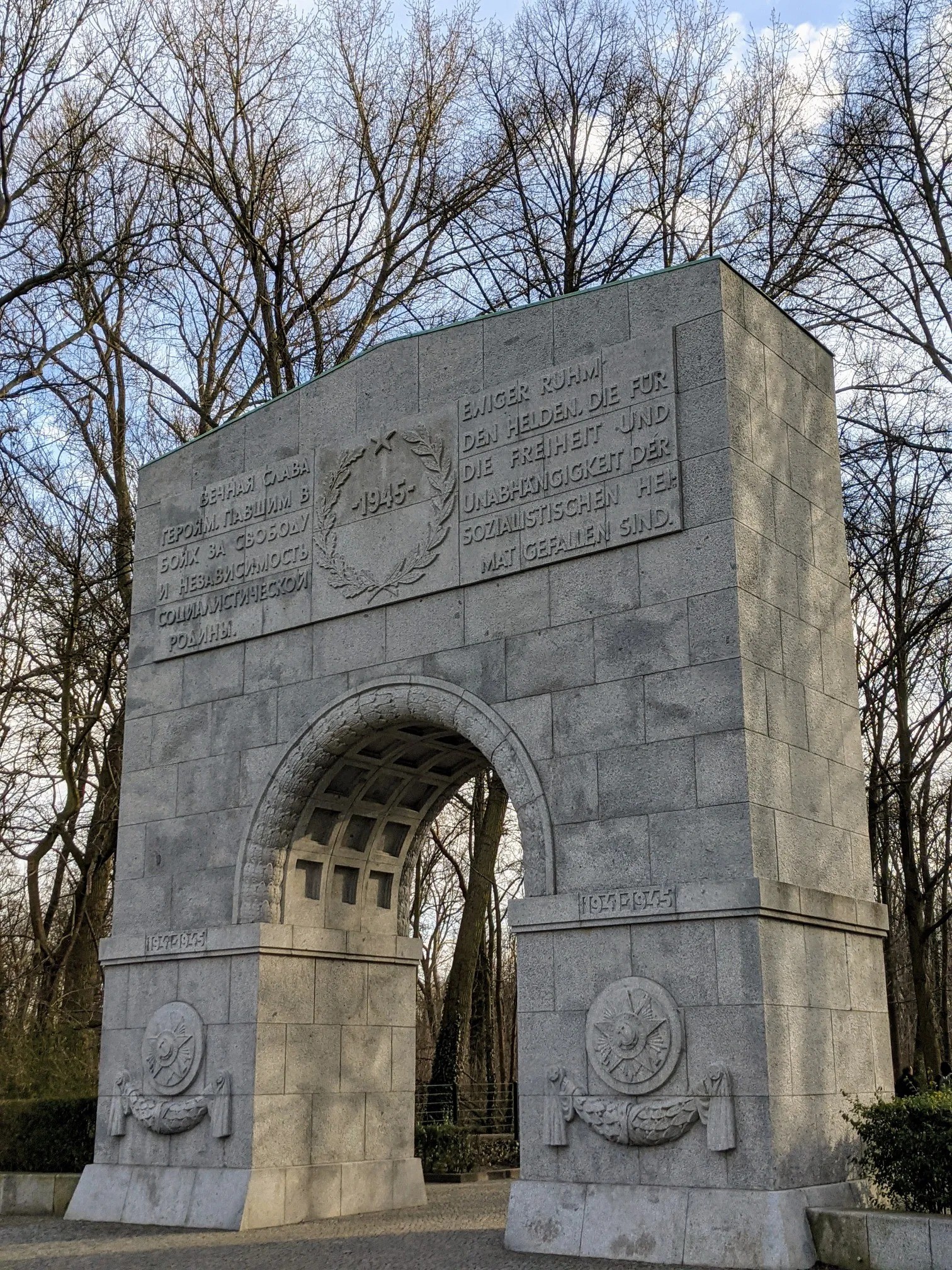 Steinerner Torbogen aus Granitquadern mit Inschriftenfeld in Deutsch und Kyrillisch, Lorbeerkranz mit Stern und 1945, dahinter kahle Bäume