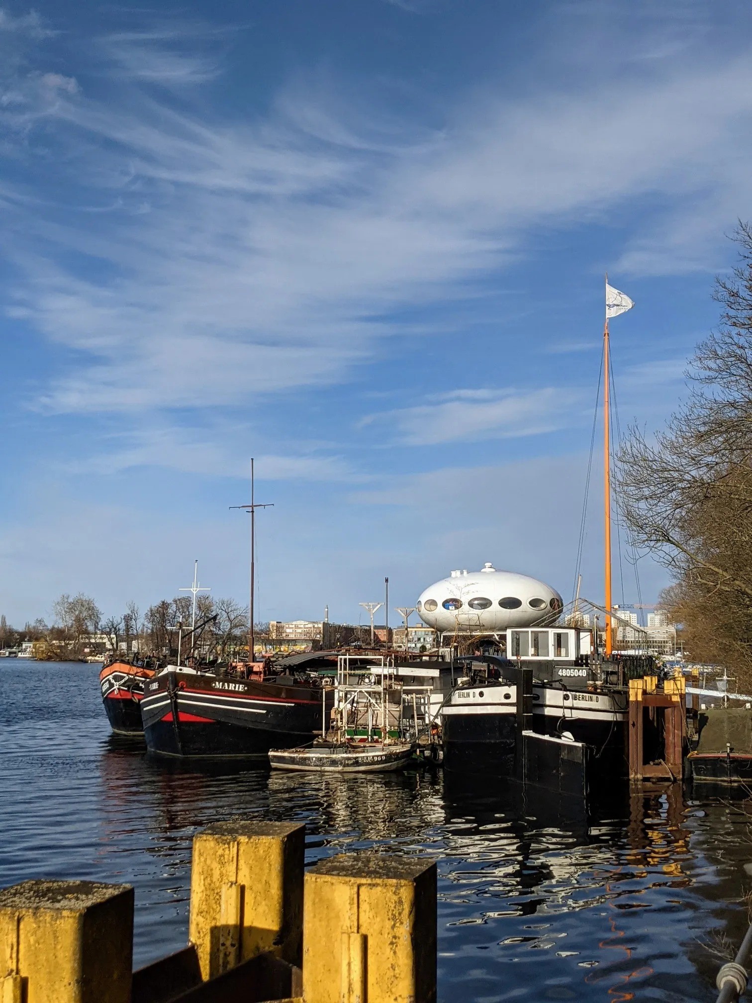 Alte Lastkähne mit den Aufschriften MARIE und BERLIN D an einem Anleger, dahinter ein weißes ovales Haus mit runden Fenstern, blauer Himmel