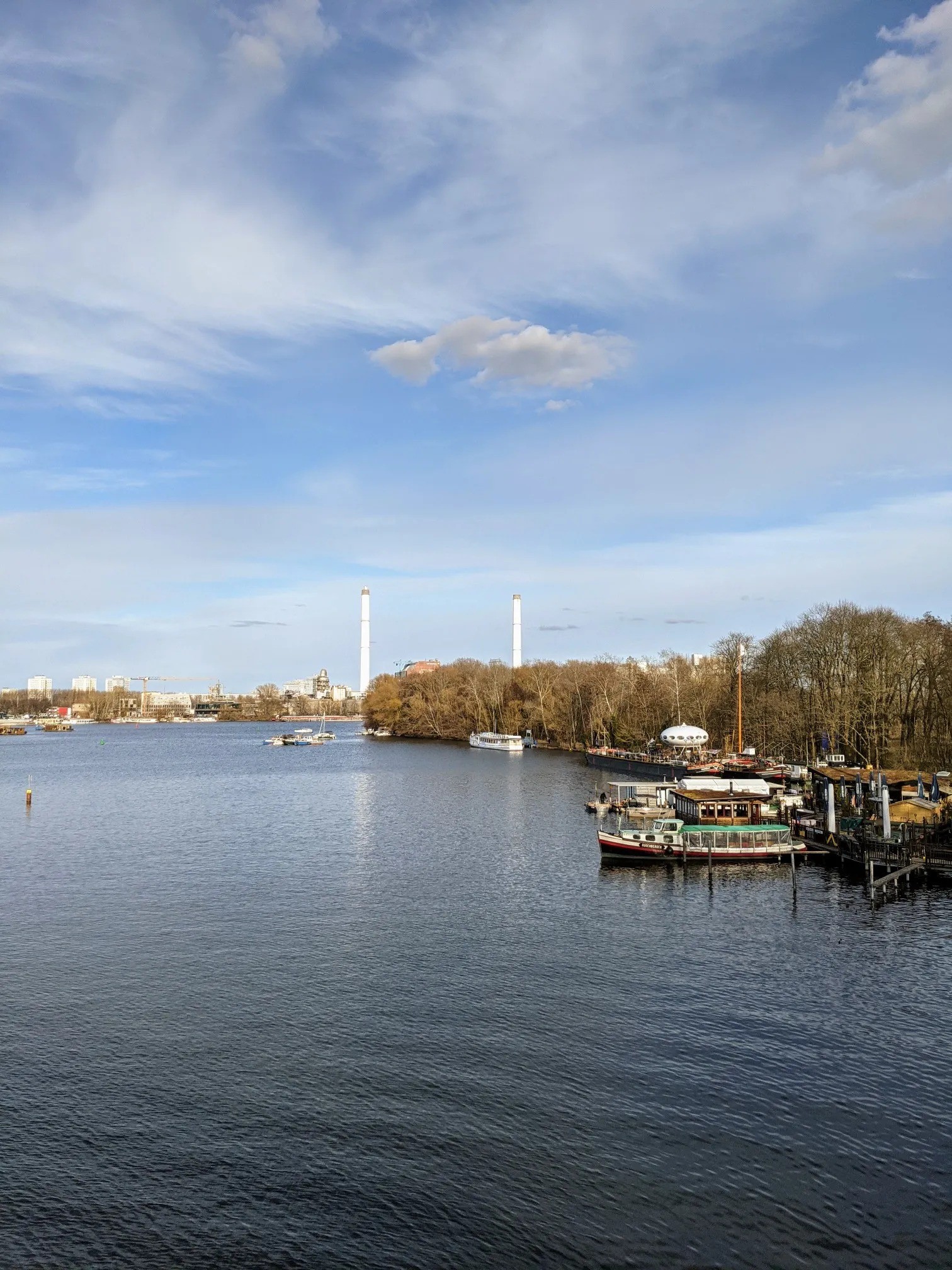 Breite Wasserfläche unter Wolkenhimmel, am Horizont zwei weiße Schornsteine, rechts am bewaldeten Ufer Boote, Stege und ein weißes ovales Haus