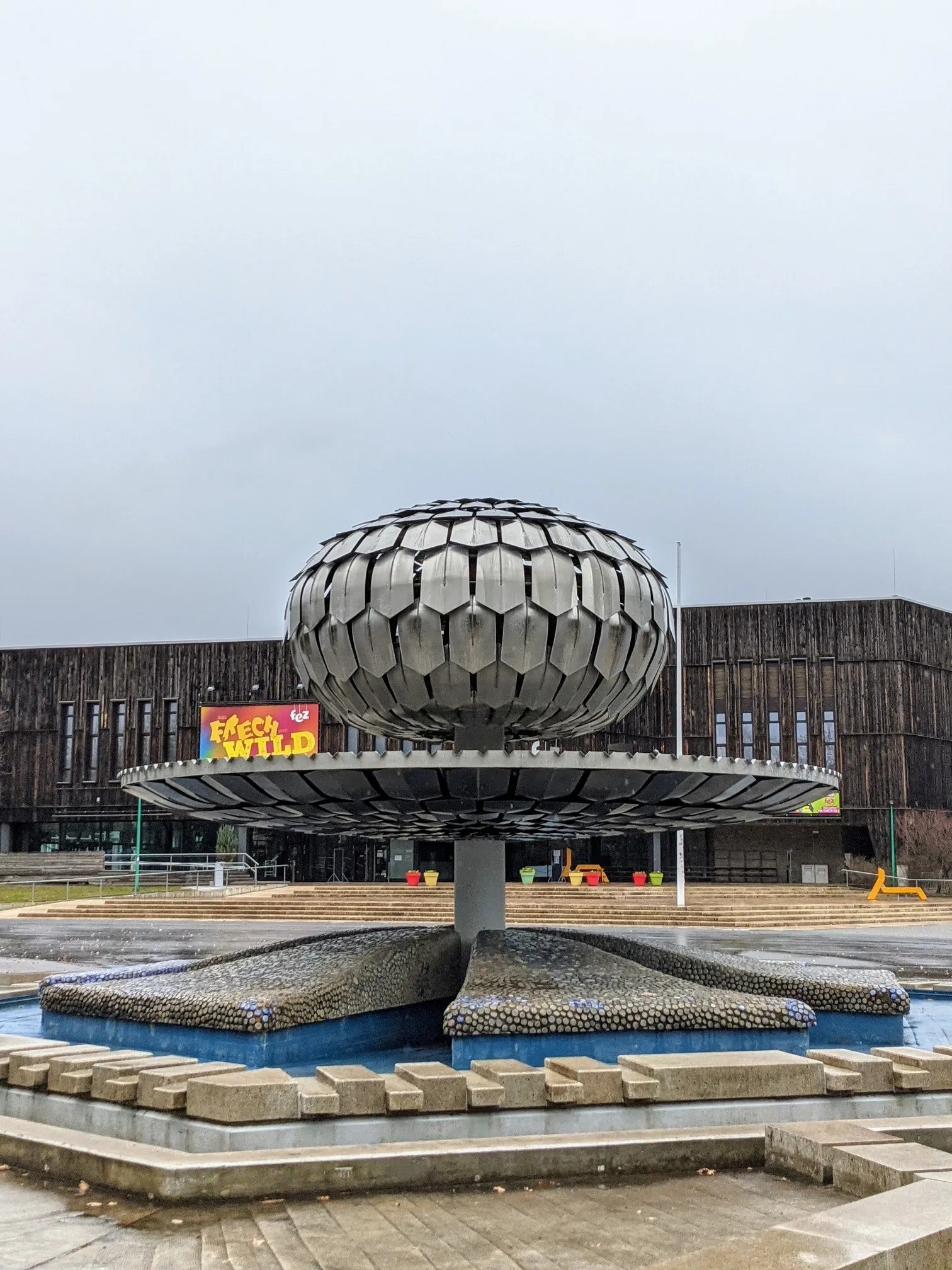 Brunnen mit einer metallenen Kugel vor einem dunklen Holzgebäude mit dem Schriftzug fez