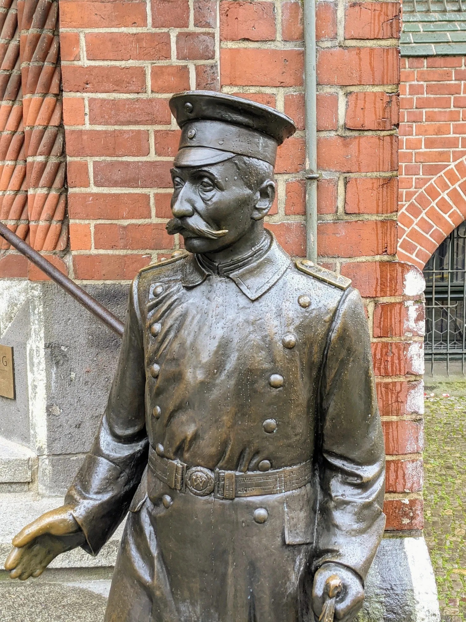 Bronzefigur eines Mannes in Uniform mit Schirmmütze vor einer Backsteinwand