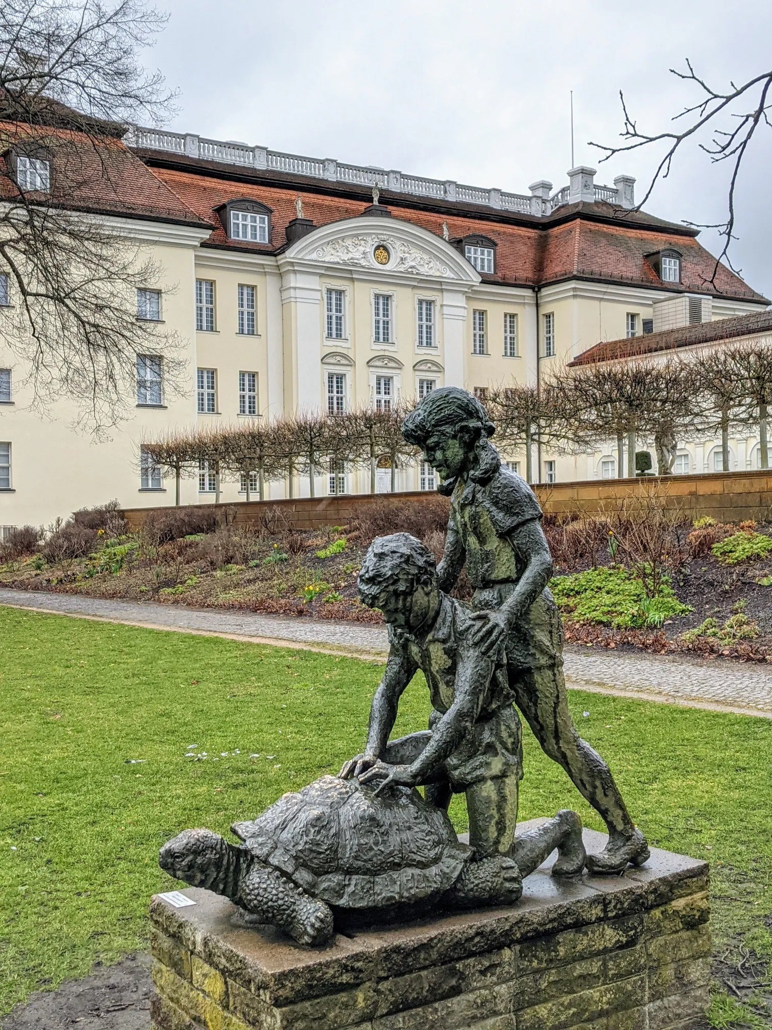 Bronzeskulptur zweier Kinder mit einer Riesenschildkröte vor einem hellen Barockschloss