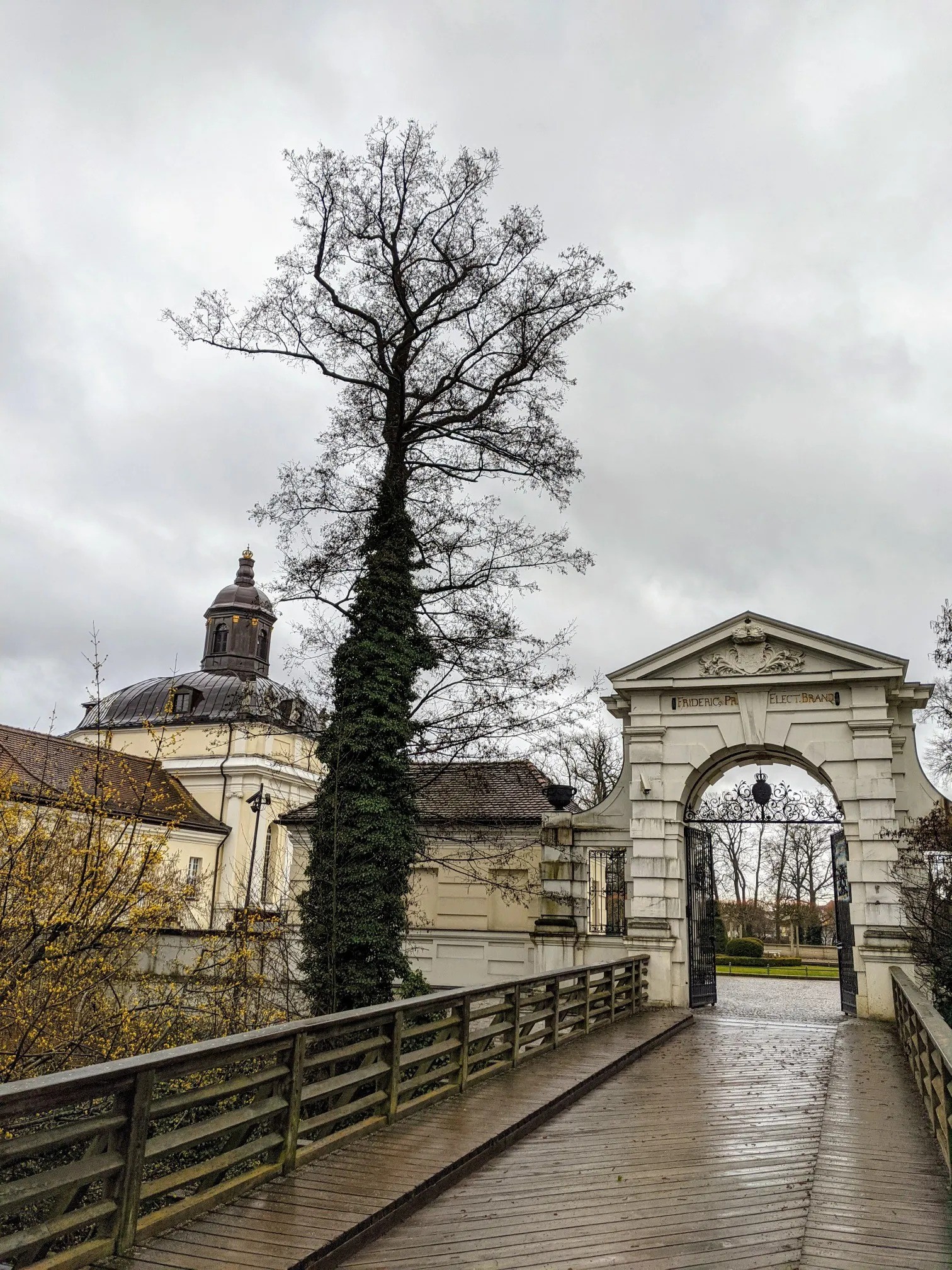 Holzbrücke zu einem weißen Torbogen mit Inschrift, dahinter die Schlosskuppel