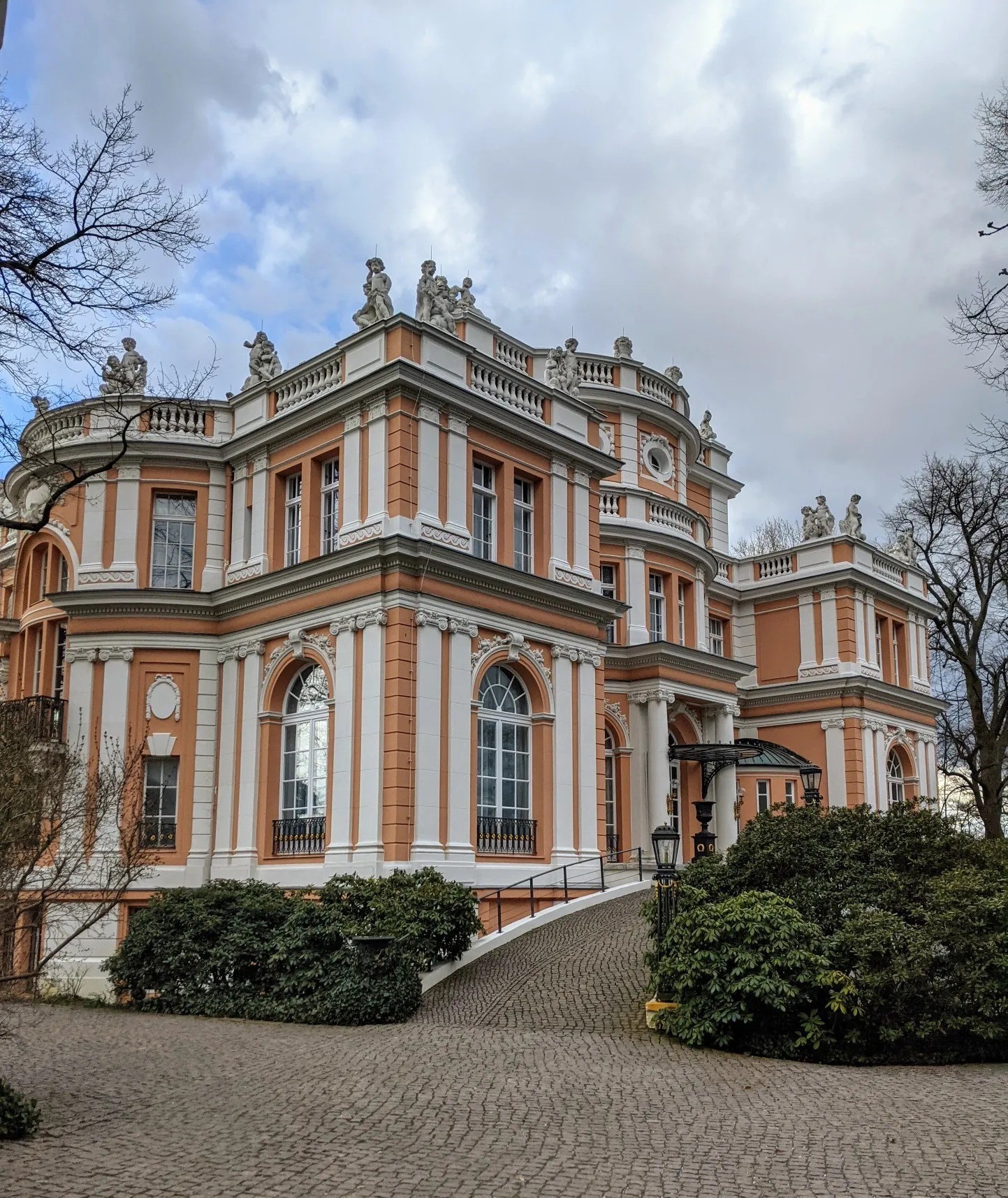 Orange-weiße Villa mit Putten und Balustraden hinter einer Kopfsteinpflasterauffahrt