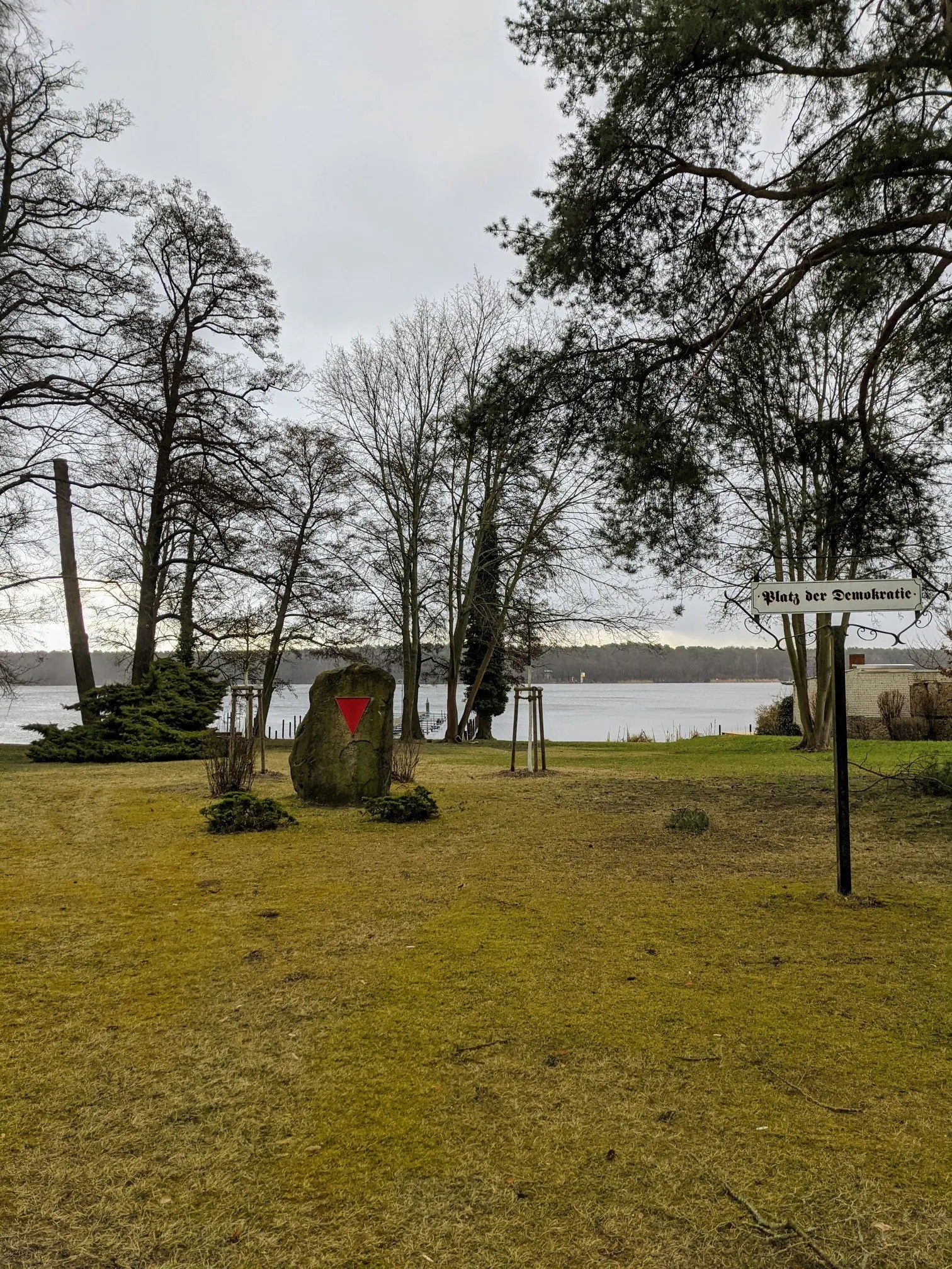 Findling mit rotem Winkel auf einer Wiese am See, rechts ein Schild Platz der Demokratie