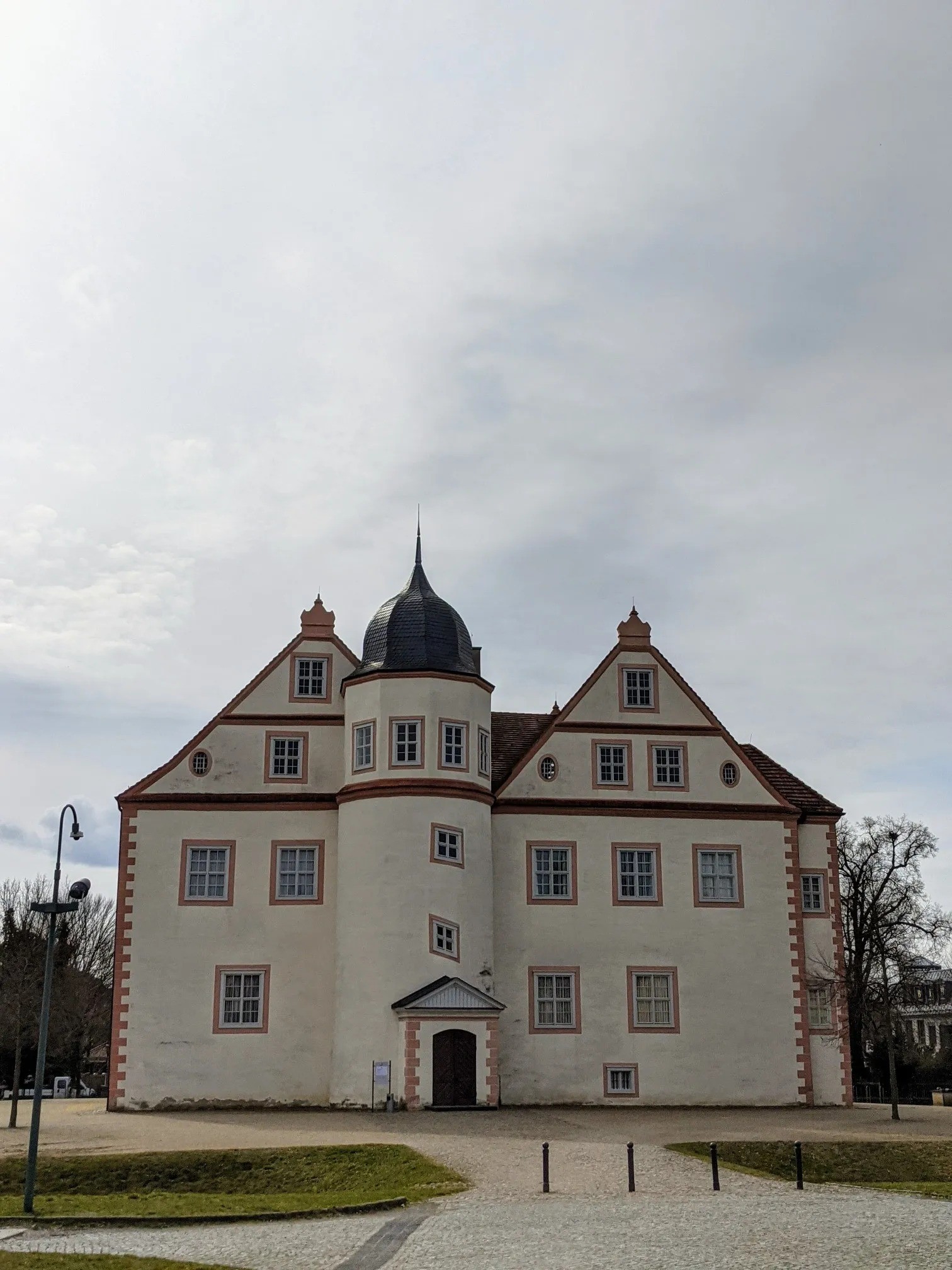 Weißes Schloss mit Rundturm und zwei Giebeln vor grauem Himmel