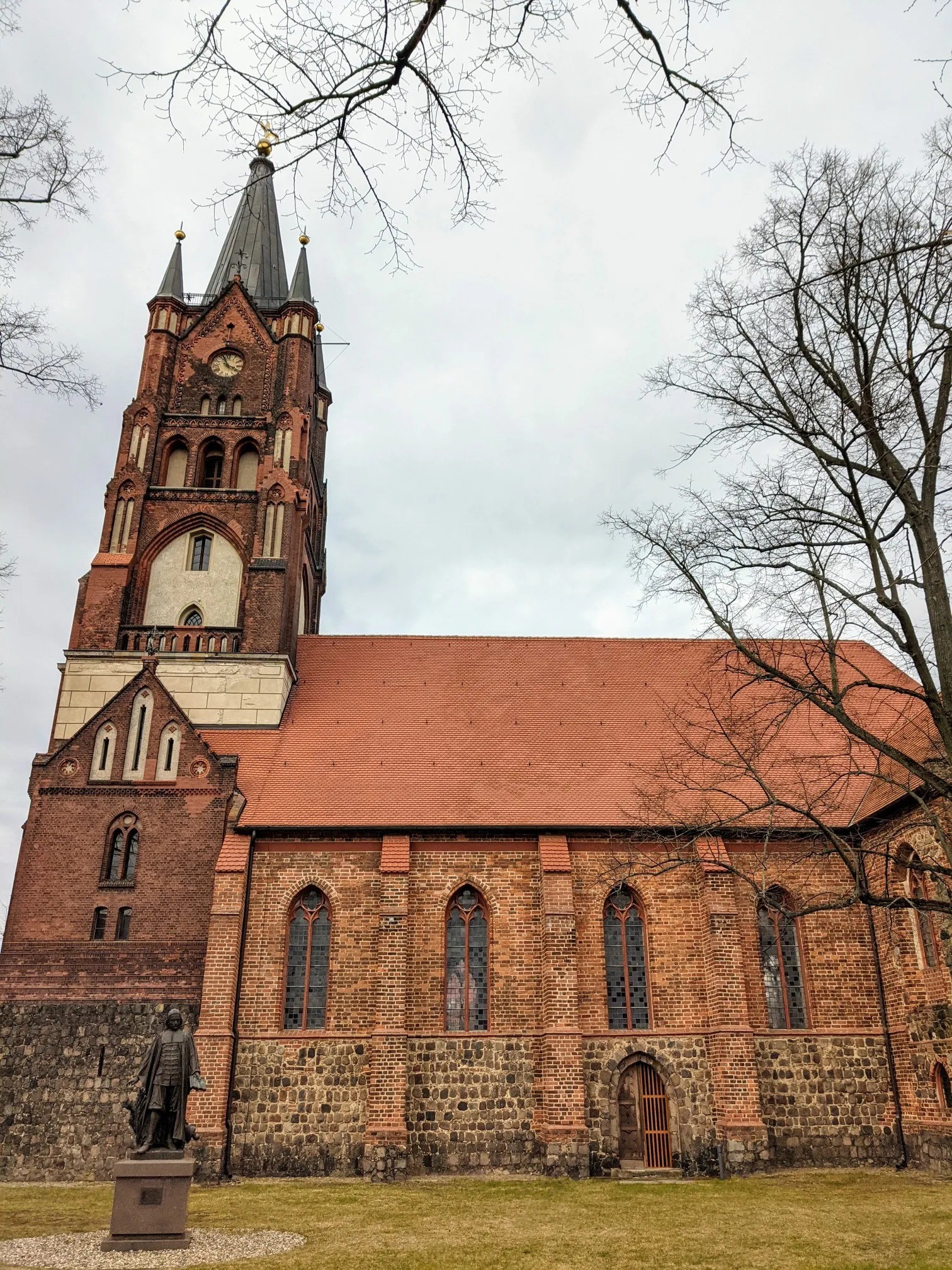 Backsteinkirche mit hohem Westturm mit Uhr und schwarzer Spitze, links unten ein Denkmal