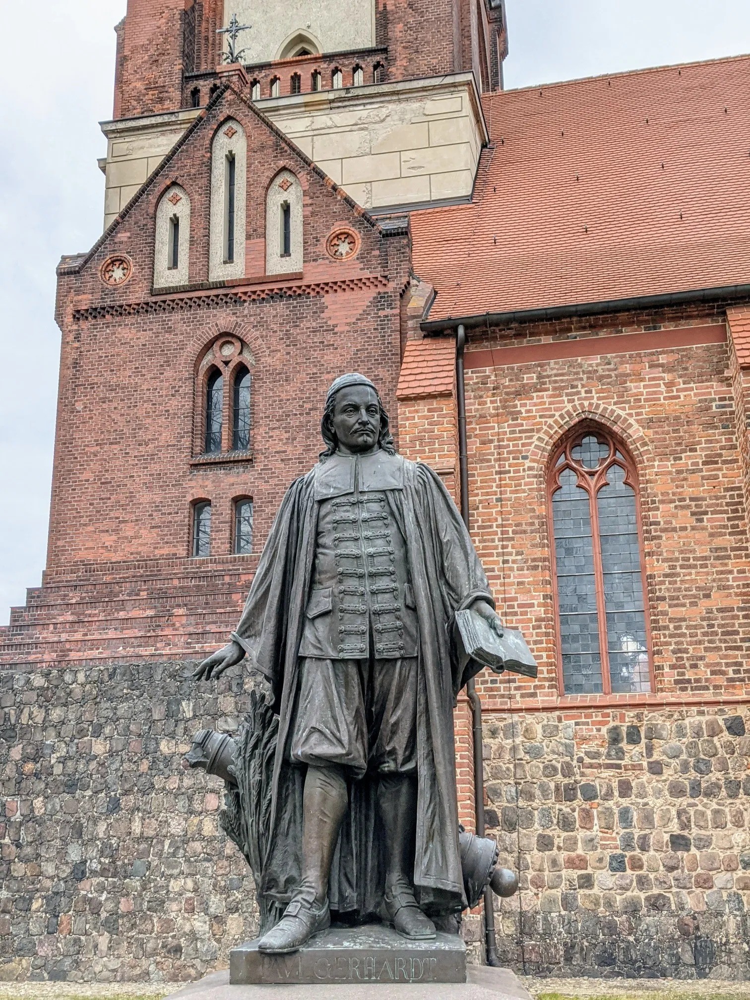 Bronzedenkmal auf einem Sockel mit der Inschrift PAUL GERHARDT, dahinter eine Backsteinkirche