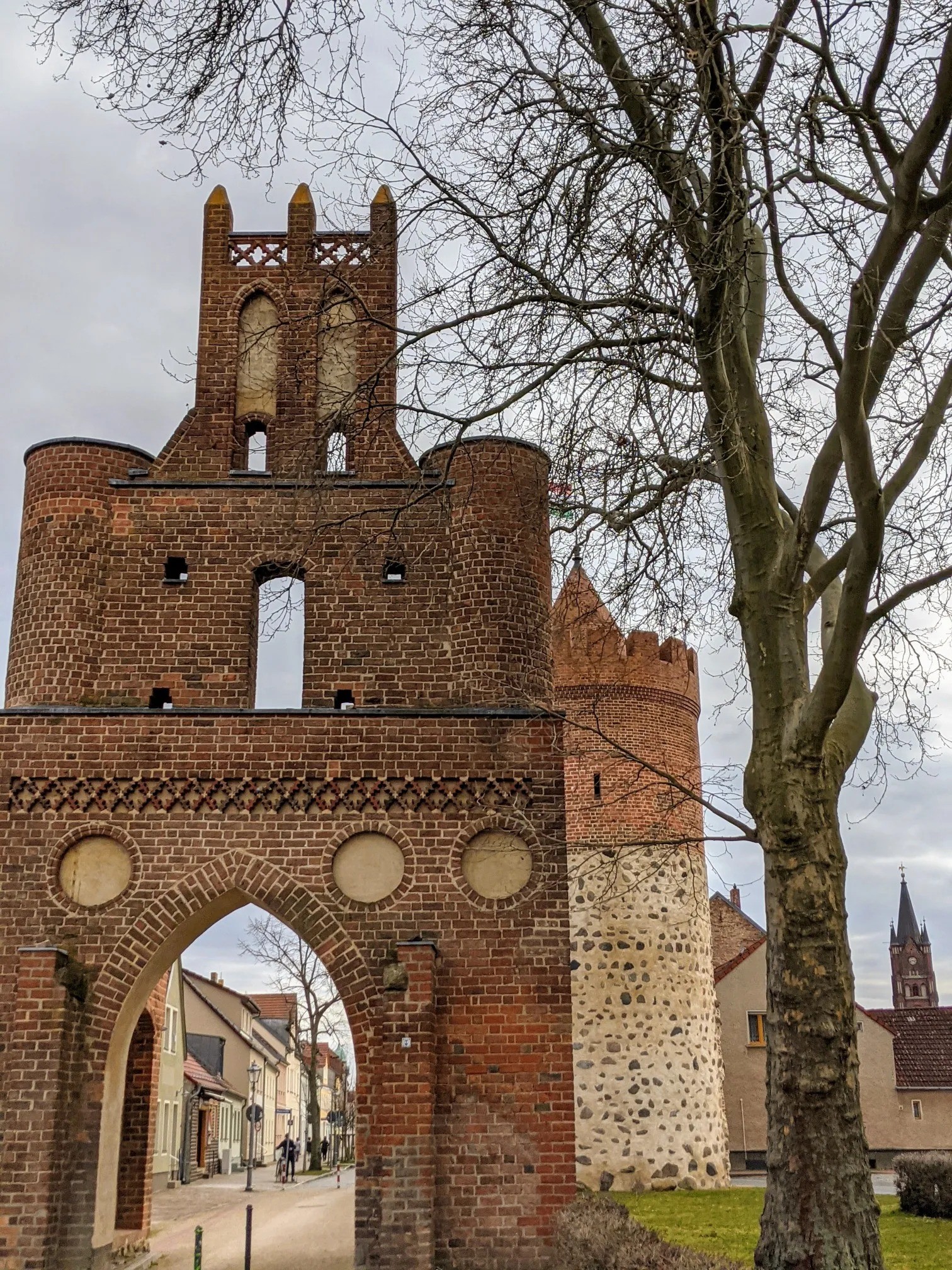 Backsteintor mit Torbogen, Rundtürmchen und Staffelgiebel, daneben ein runder Wehrturm, durch den Bogen eine Gasse mit Radfahrer