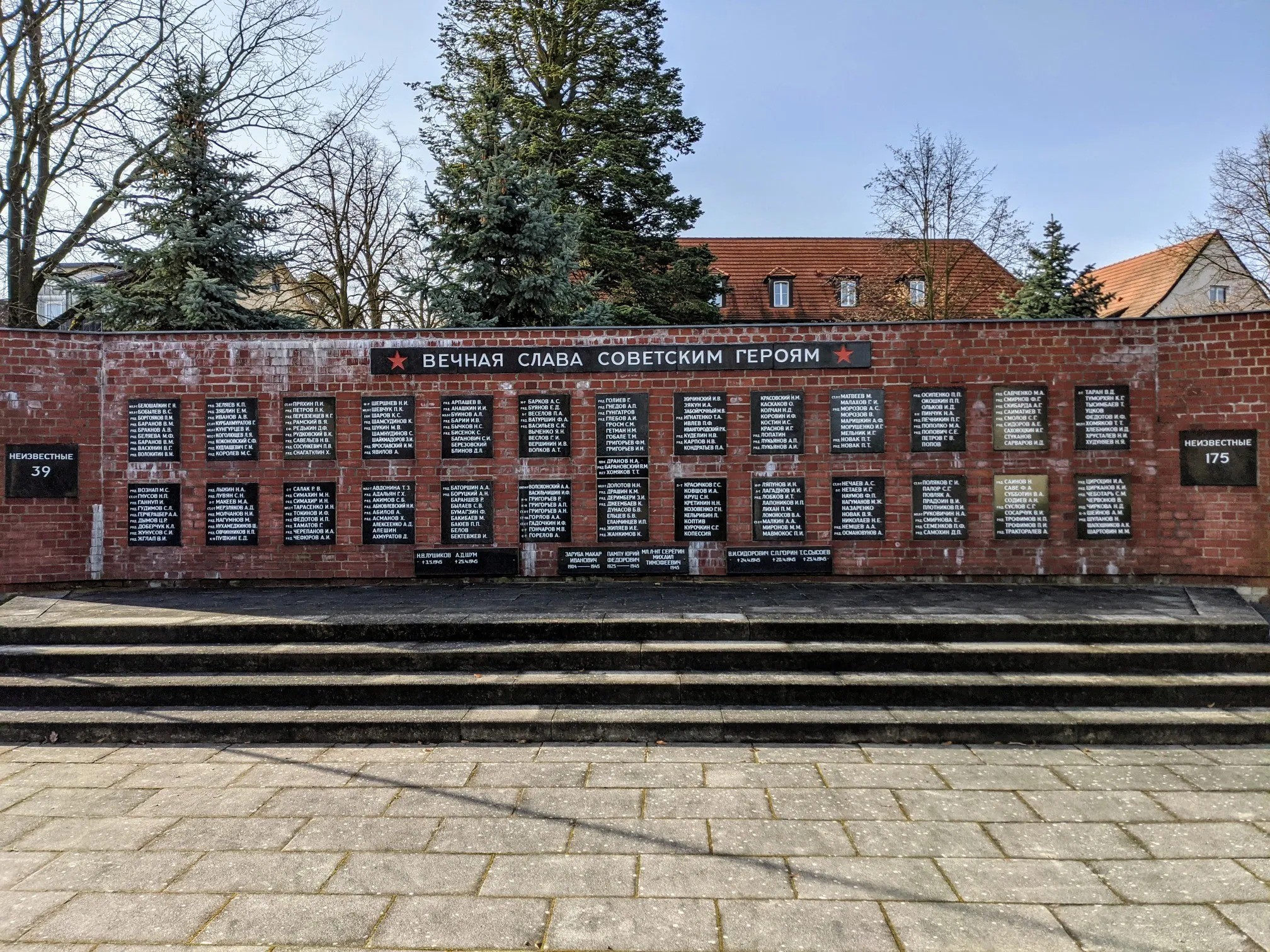 Rote Backsteinwand mit schwarzen Namenstafeln und kyrillischer Inschrift, Stufen davor