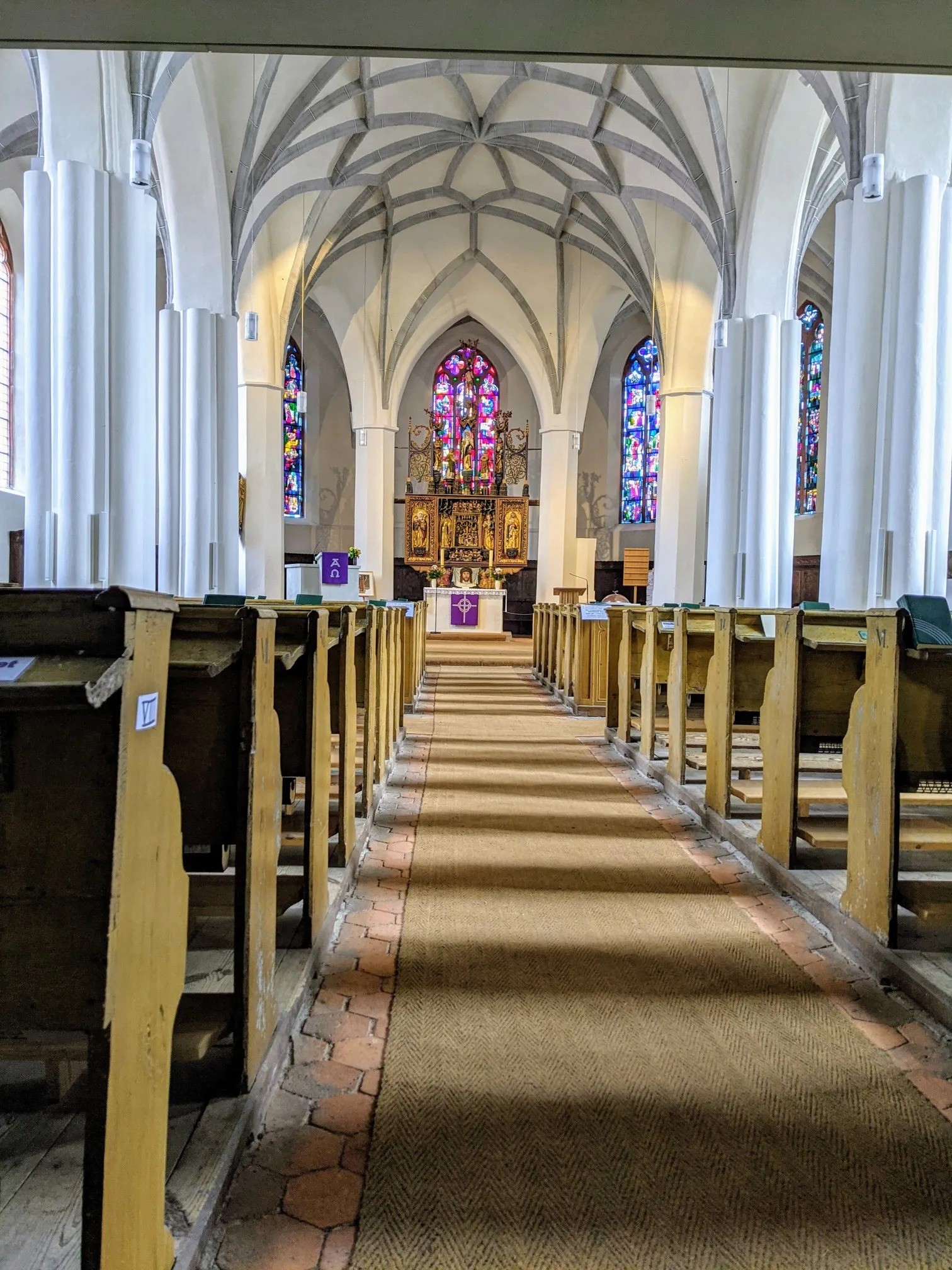 Kirchenschiff mit weißen Säulen, Netzgewölbe, Altar und bunten Glasfenstern