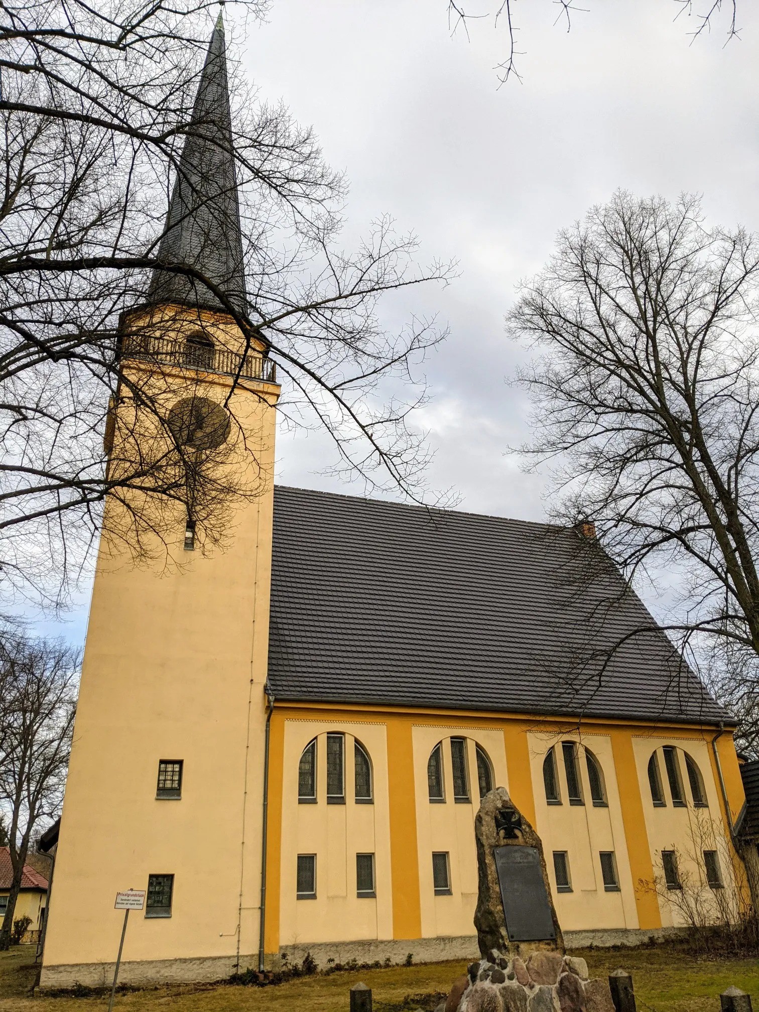 Gelbe Kirche mit spitzem Turm und einem Gedenkstein davor