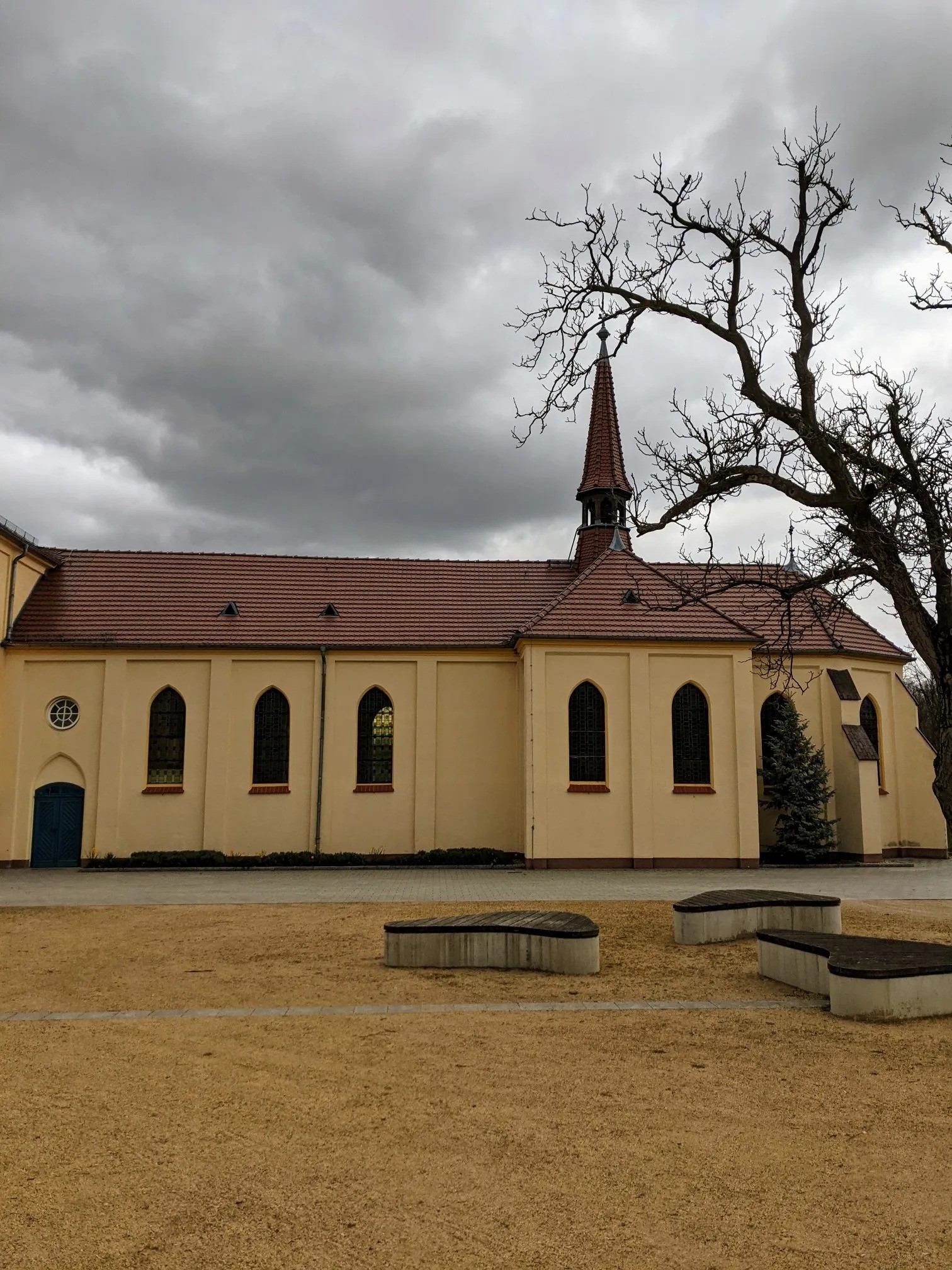 Gelbe Kirche mit Dachreiter hinter einem Sandplatz mit Bänken