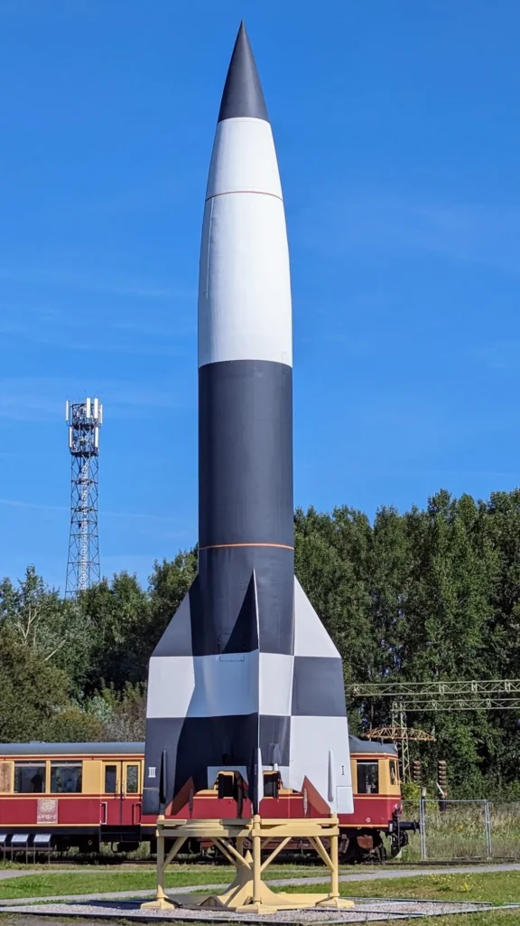 Schwarz-weißes Modell der Rakete Aggregat 4 auf dem Freigelände, dahinter ein rot-gelber Triebwagen der Werkbahn