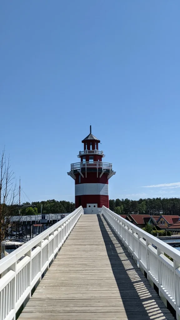 Holzsteg mit weißem Geländer zum rot-weißen Leuchtturm im Hafendorf Rheinsberg