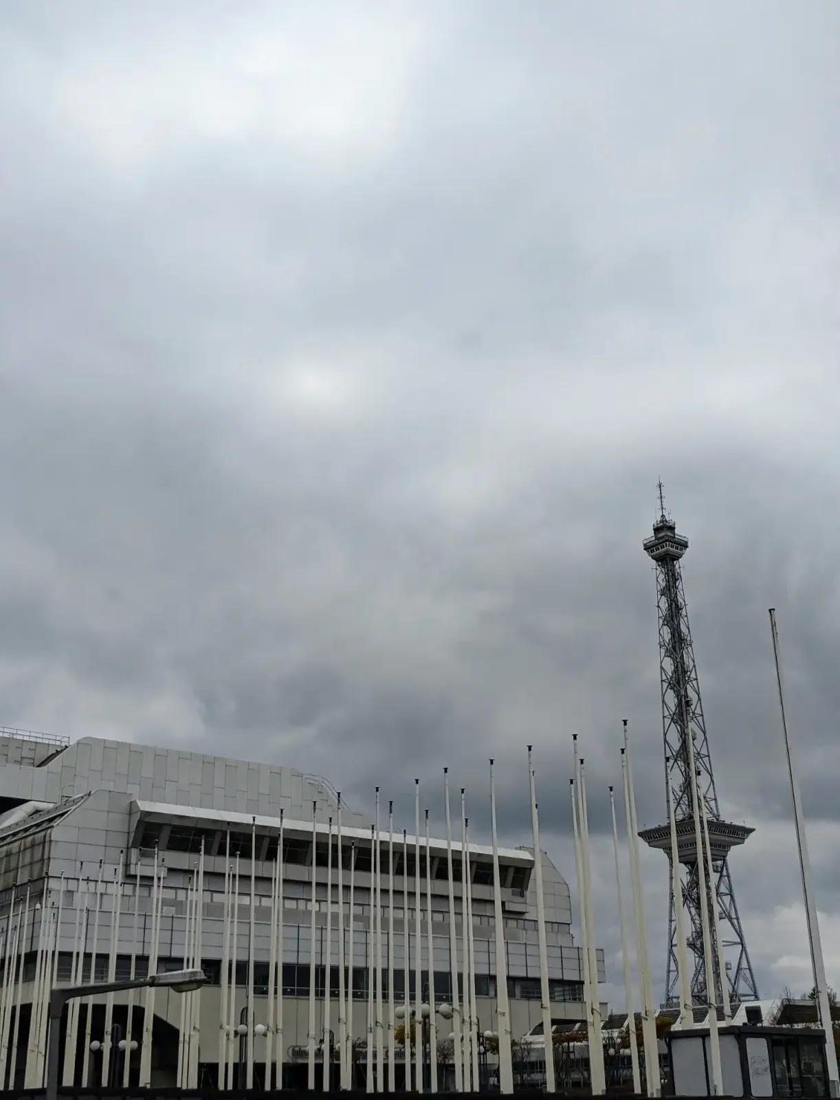 Grauer Hallenbau des Berliner Messegeländes mit einer Reihe Fahnenmasten, dahinter der Funkturm