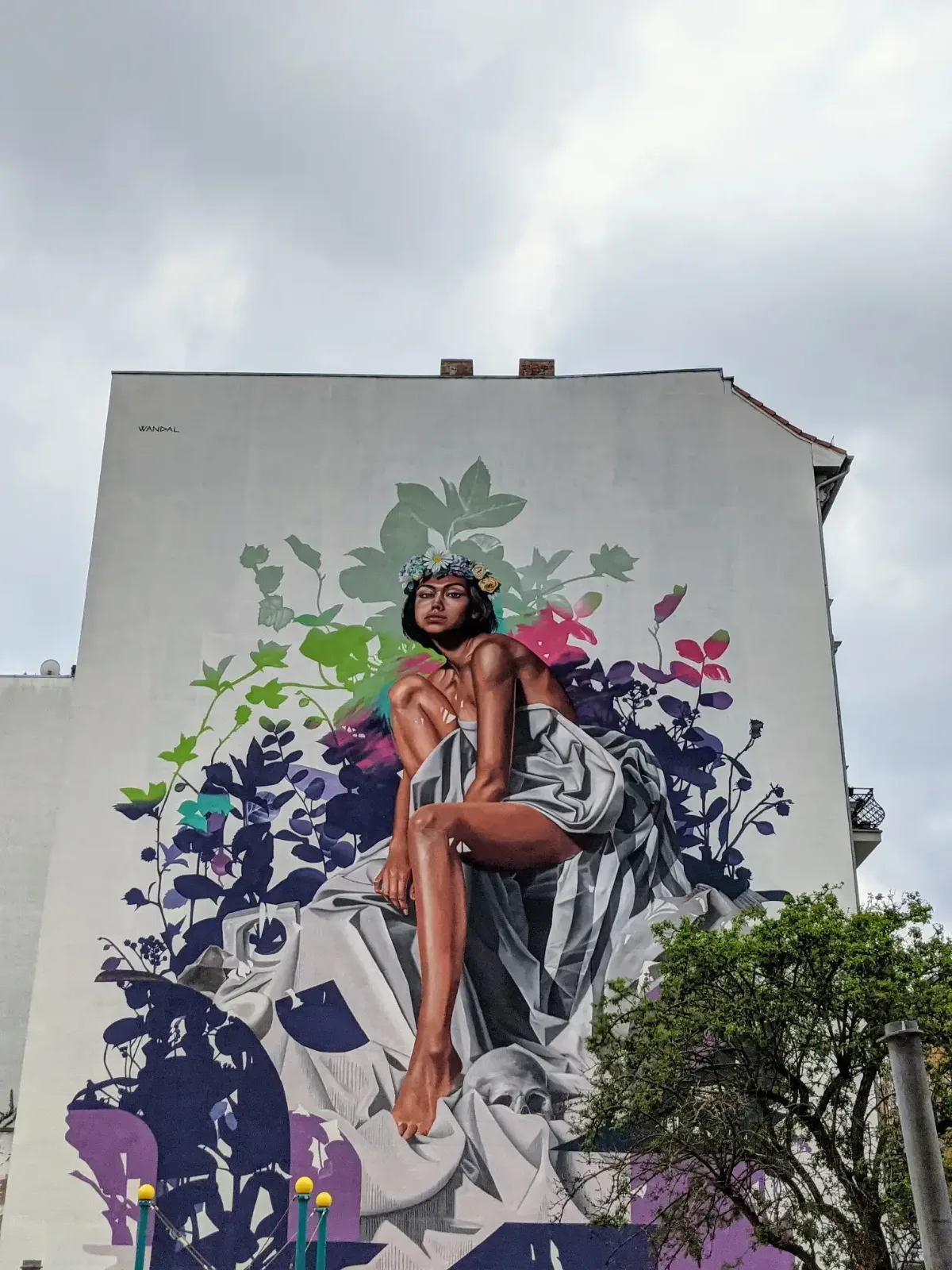 Wandbild an einer weißen Brandwand in Berlin-Moabit: sitzende Frau im Tuch vor Blättern und Blüten in Violett und Grün
