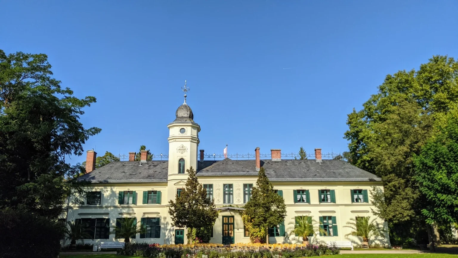 Gartenseite von Schloss Britz mit Turm und grünen Fensterläden unter blauem Himmel