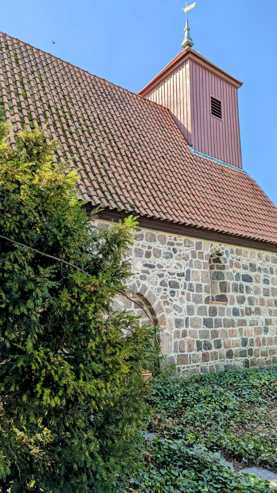 Feldsteinkirche mit Satteldach und verbrettertem Fachwerkturm, davor ein Nadelbaum, an der Breiten Straße