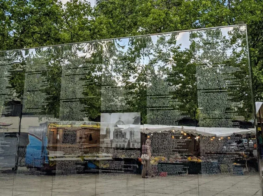 Spiegelwand auf dem Hermann-Ehlers-Platz in Berlin-Steglitz: Namenslisten auf poliertem Stahl, darin gespiegelt Bäume und Marktstände